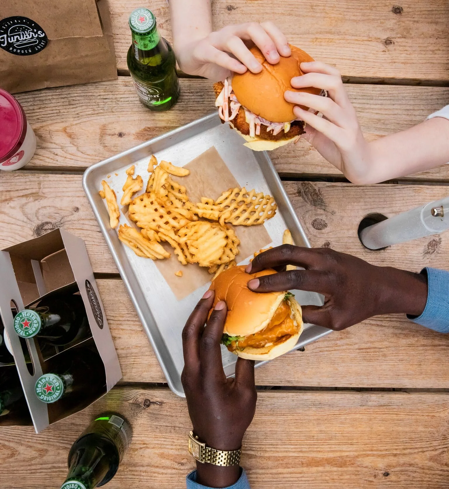 Two people holding burgers above a tray of waffle fries with several beer bottles on a wooden table.