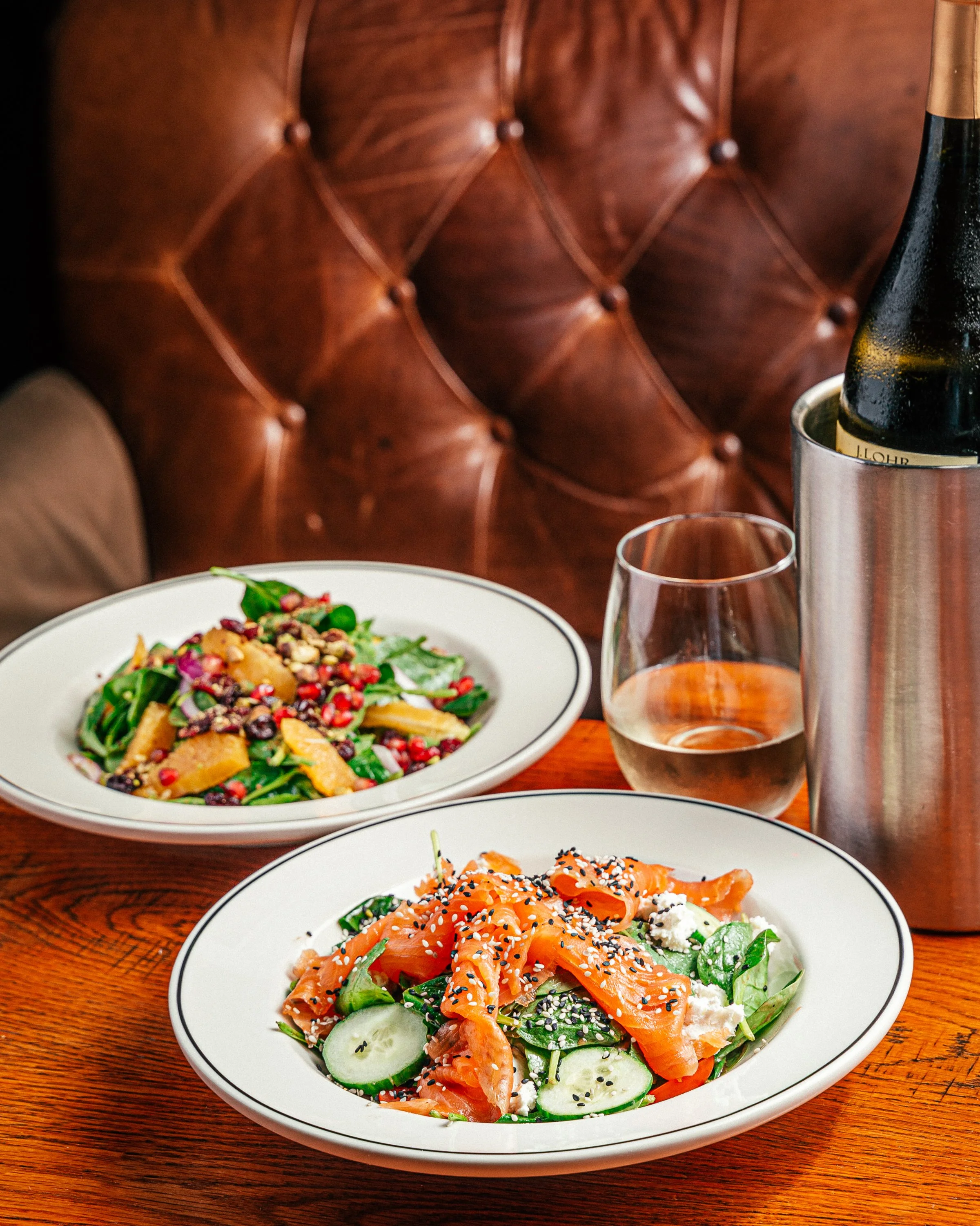 Two plates of salad on a wooden table, one with smoked salmon, cucumbers, and sesame seeds; the other with greens, pomegranate seeds, and cheese. A glass of rosé wine, a bottle in an ice bucket, and a brown leather tufted seat in the background.