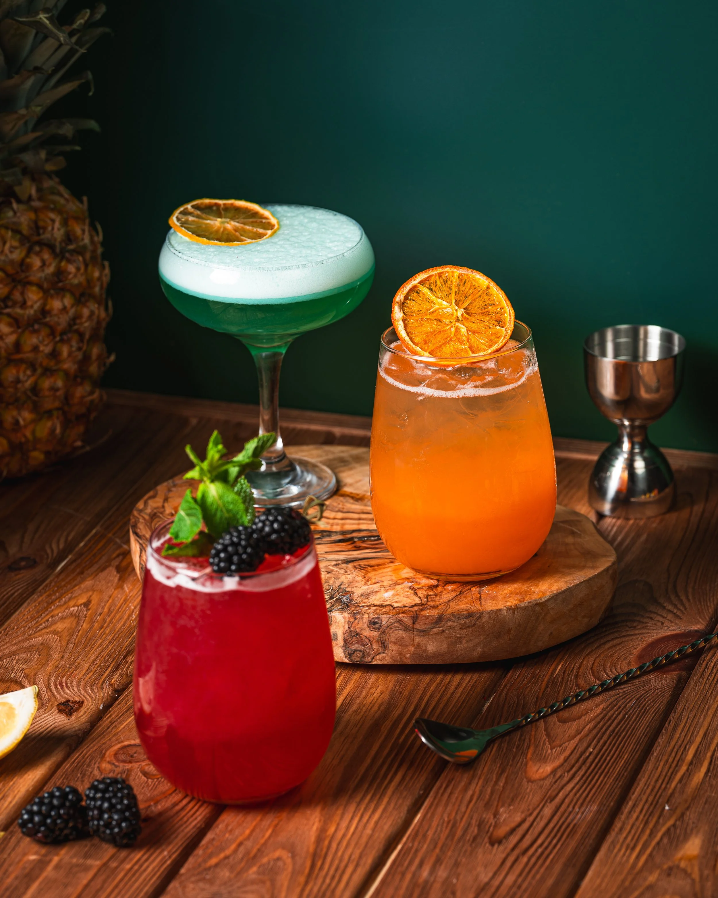 Three colorful cocktails with fruit garnishes on a rustic wooden table, with a pineapple partially visible in the background.