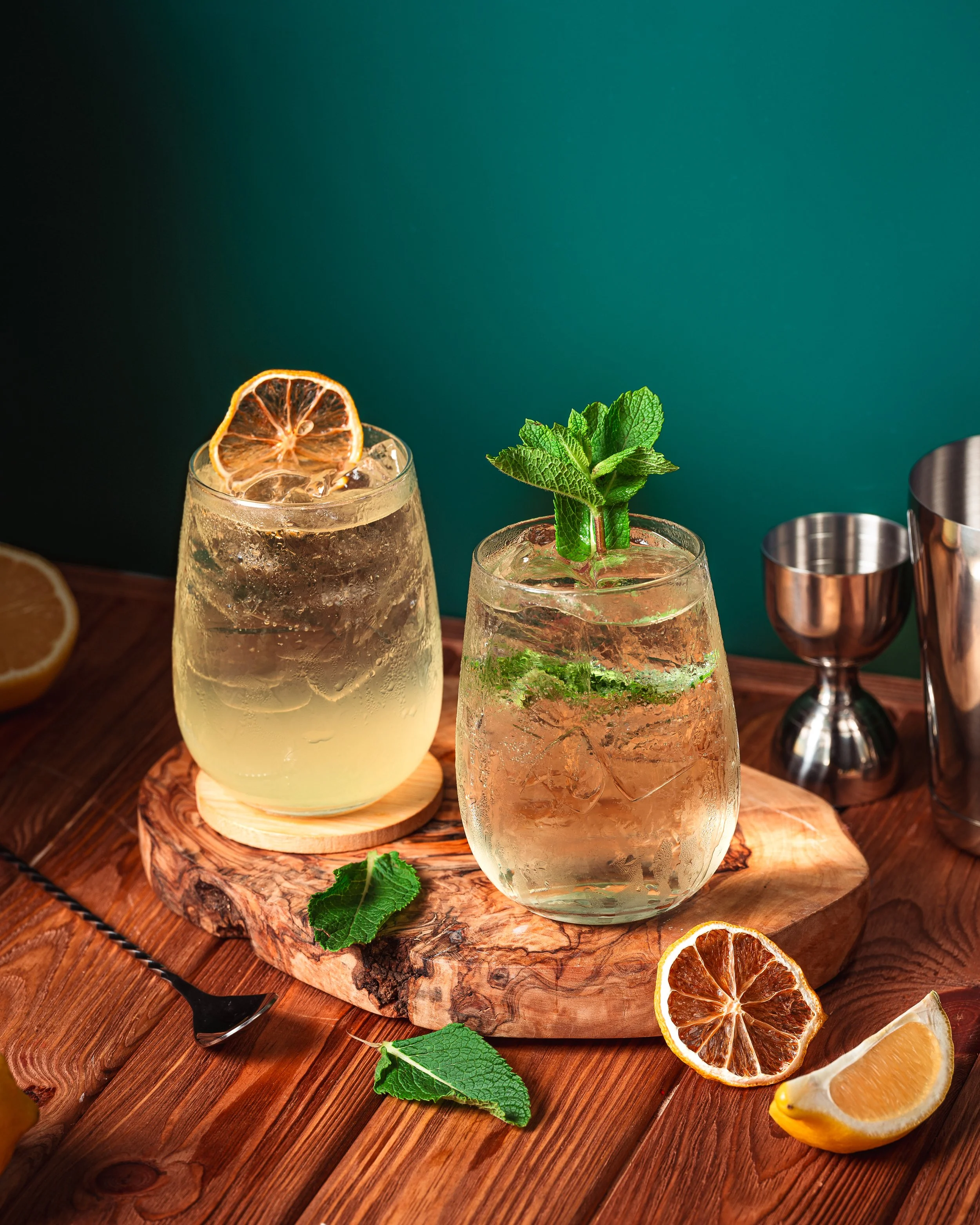 Two glasses of citrus cocktail with mint and dried orange slices, on a wooden board with lemon slices and mint leaves, against a dark teal background.