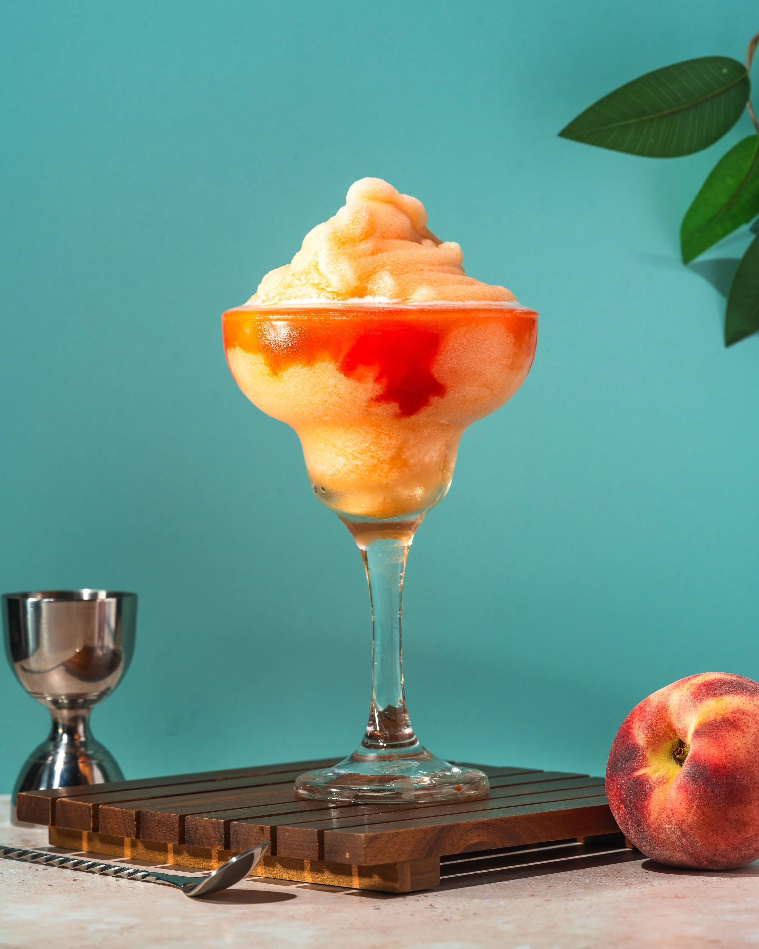 Peach and red syrup frozen dessert served in a large dessert glass with a stem, on a wooden serving board, with a whole peach, a small metal jigger, and a cocktail spoon nearby, against a teal background with green leaves.