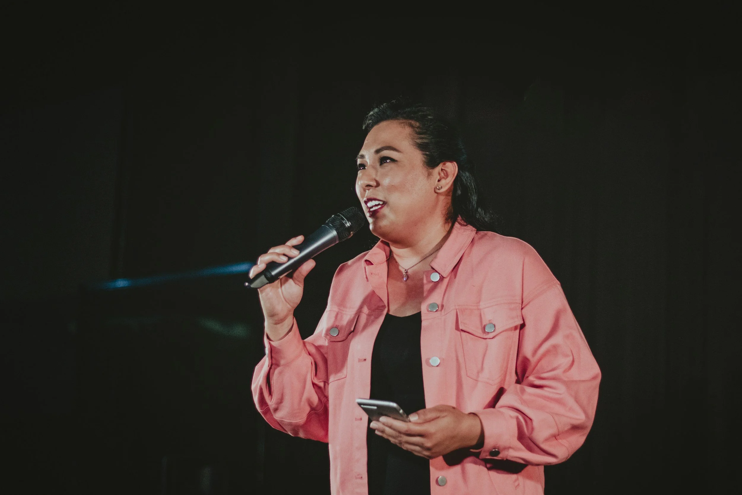 Woman speaking into microphone, holding phone, wearing pink jacket and black shirt, in dark setting.