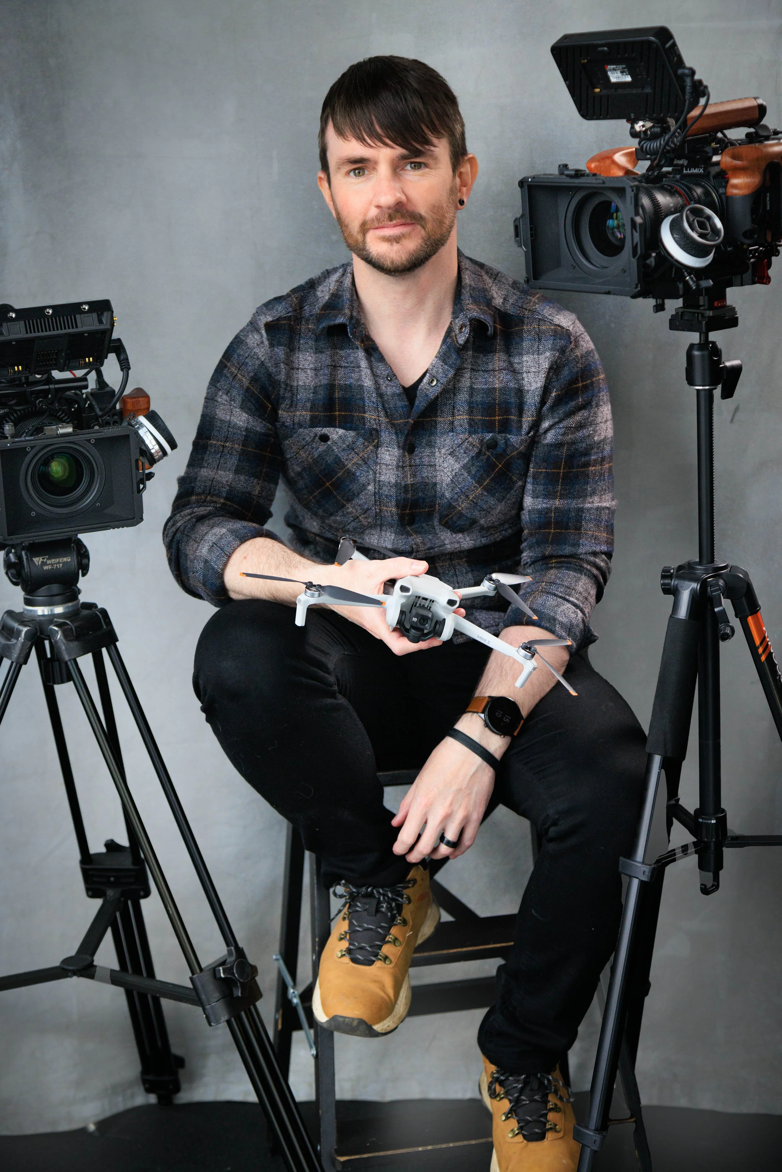 A man with dark hair and a beard sitting on a chair in front of a plain gray wall, holding a drone, with professional filming equipment including a camera and lighting setup around him.