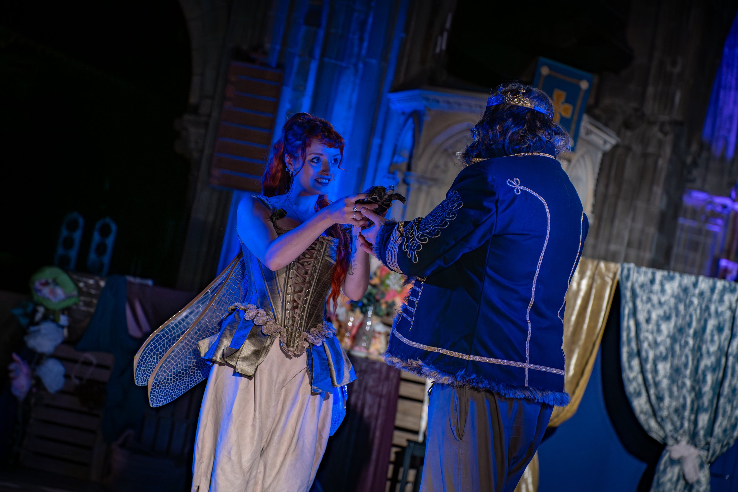 Two people dressed in elaborate costumes, one with red hair and the other with a crown, exchanging a small object in a dimly lit setting with medieval-style stone architecture and blue lighting.