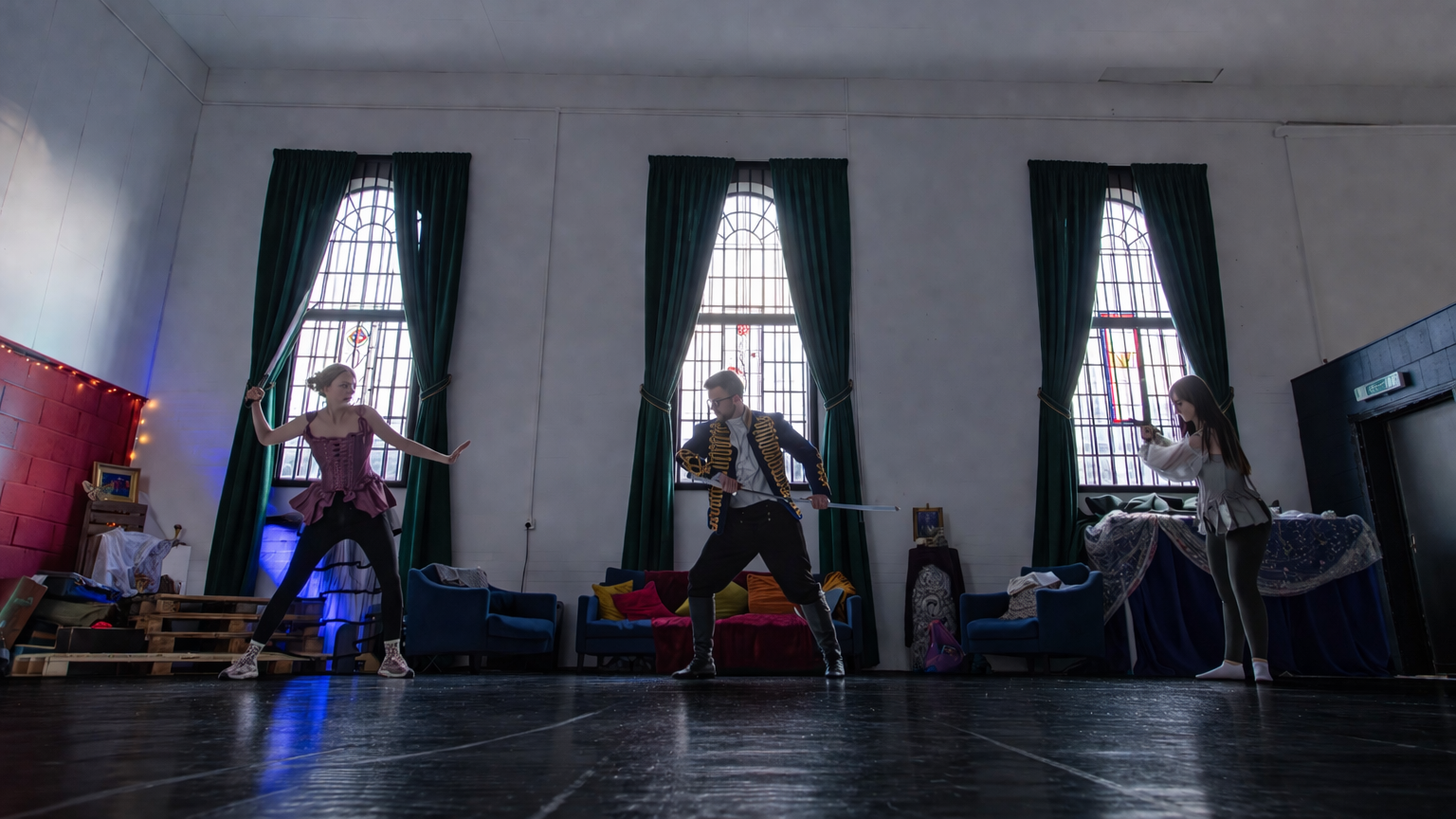 Three people practicing sword fighting in a room with large stained glass windows, blue couches, and various items on the floor.