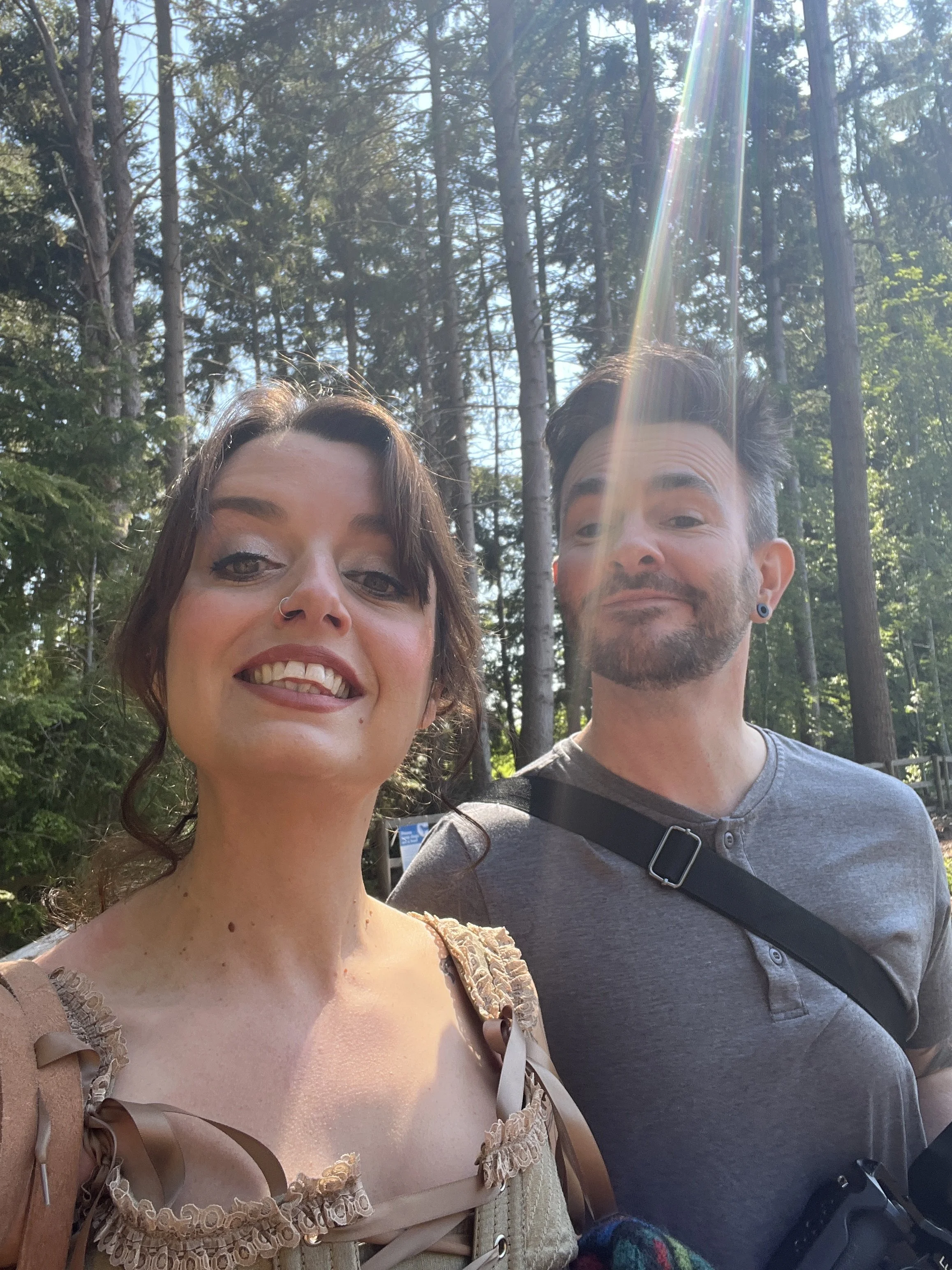 A woman and a man are taking a selfie outdoors in a forested area with tall trees in the background and sunlight filtering through the trees.