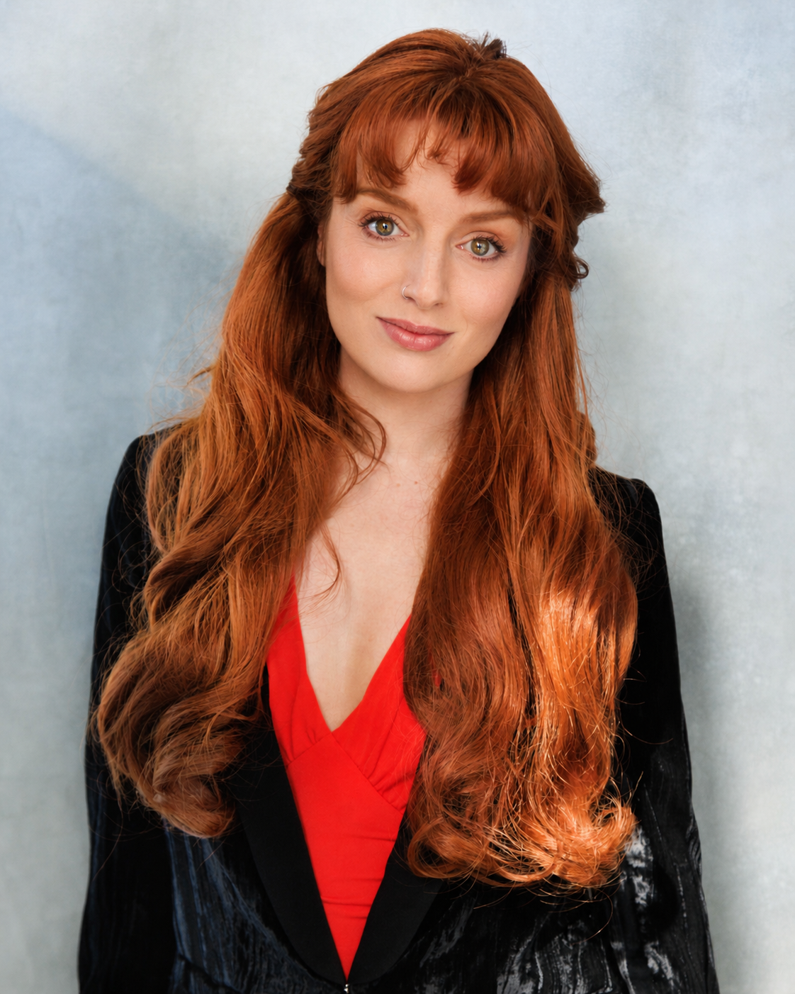 A woman with long, wavy red hair and a nose ring, wearing a black blazer and red top, smiling slightly against a light-colored background.