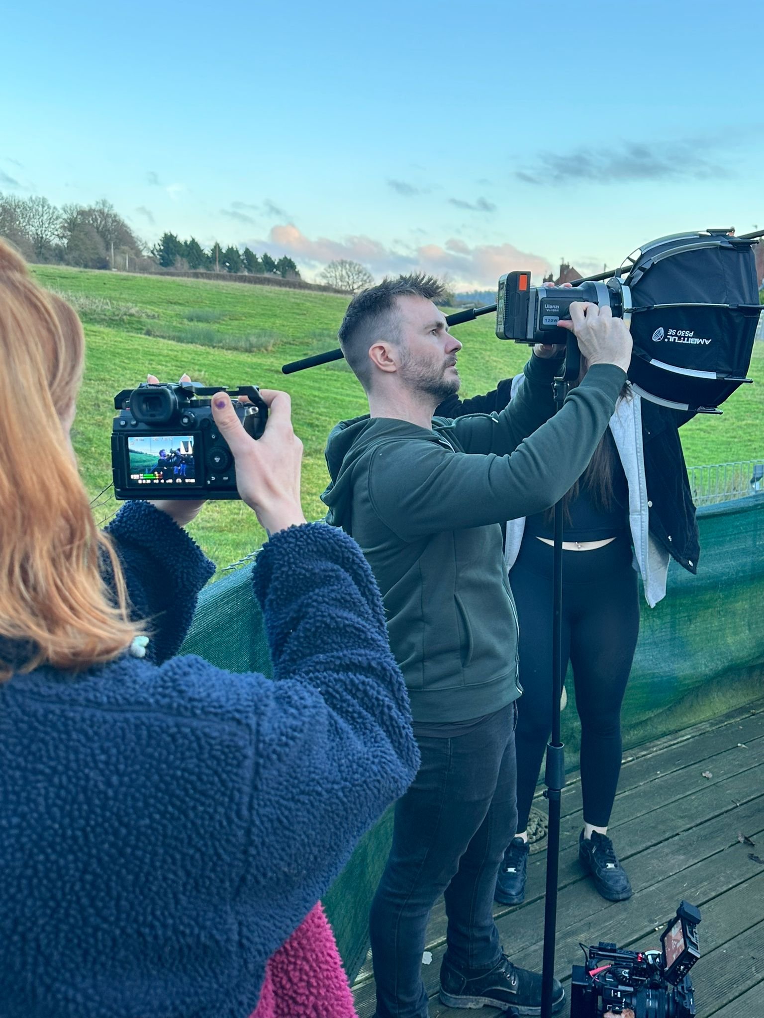 A film crew outdoors on a wooden deck filming a scene. One person is holding a professional camera, another with a large camera mounted on a stabilizer, and a third filming with a handheld camera. The background features green fields and a cloudy sky