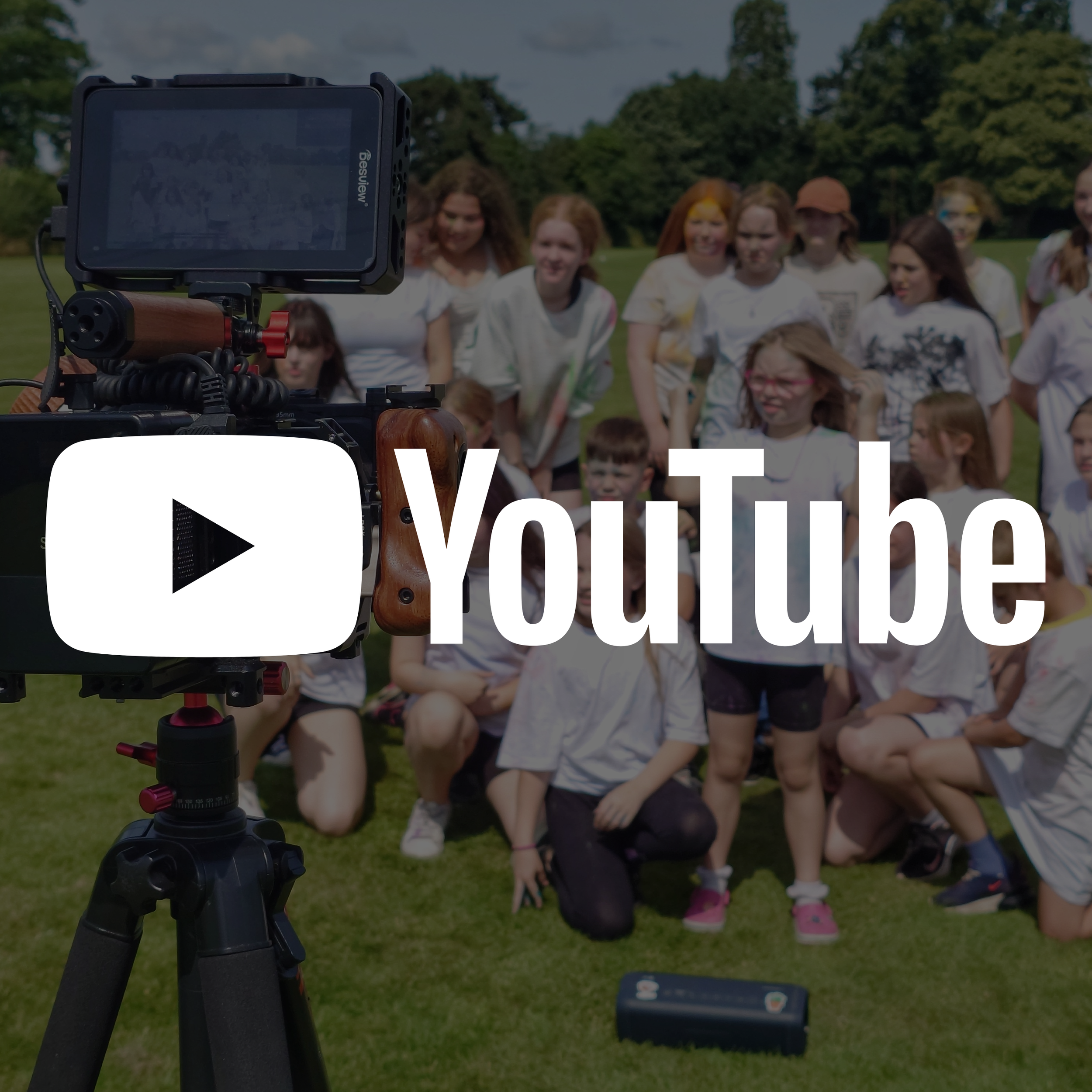 A camera on a tripod taking a photo of a group of children outdoors on a grassy field, with trees and a cloudy sky in the background.