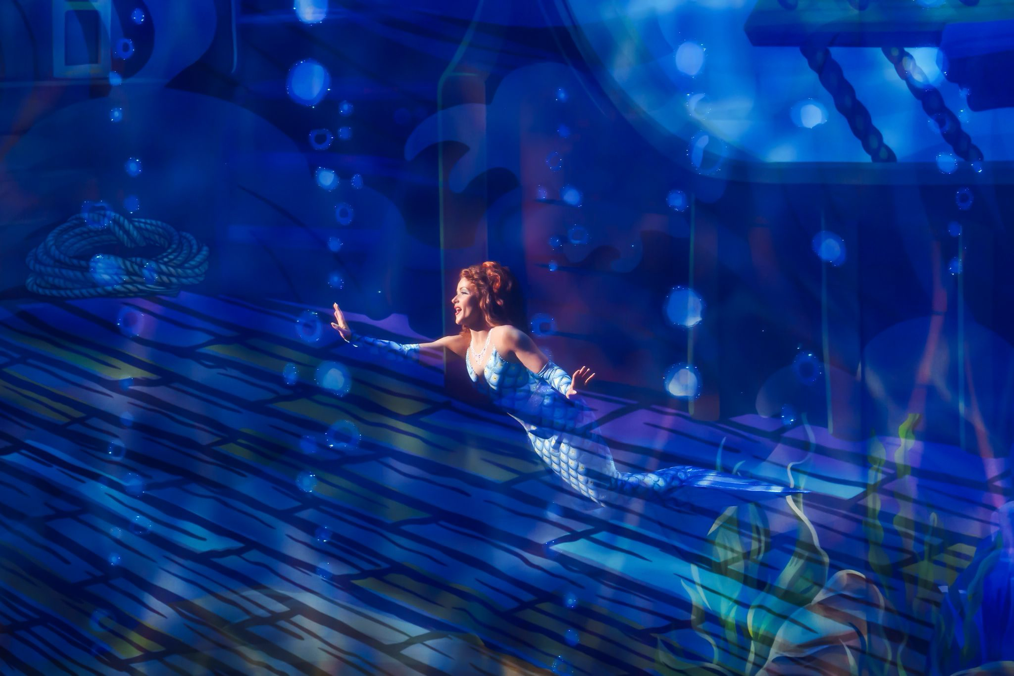A woman with red hair and a shiny, blue, mermaid costume is performing on a stage illuminated with blue lighting, with a background of underwater-themed decorations and projections.