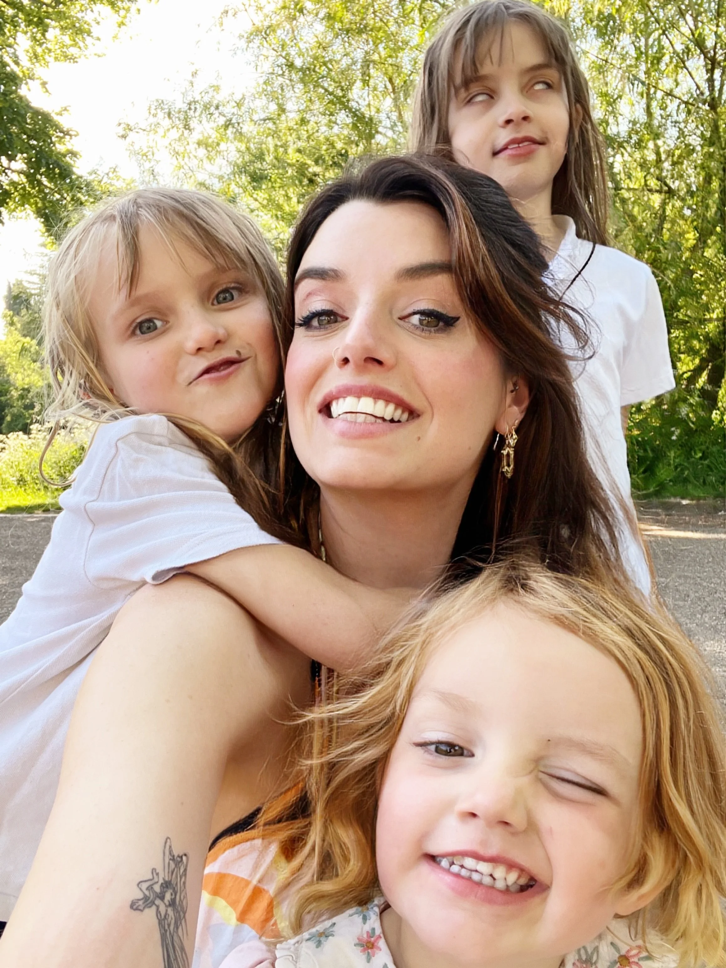 A smiling woman with dark hair and earrings taking a selfie with two young children outdoors among green trees, one girl with light brown hair and another with red hair, both smiling.