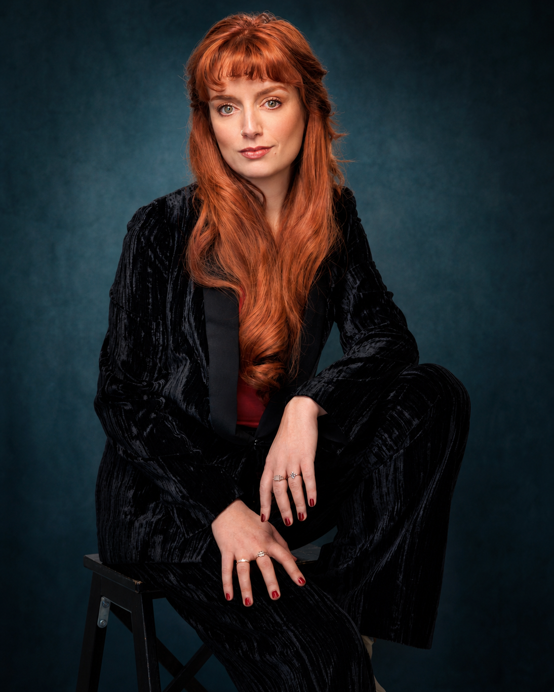 A woman with long red hair and a nose piercing, wearing a black velvet suit, sitting on a stool against a dark teal background.