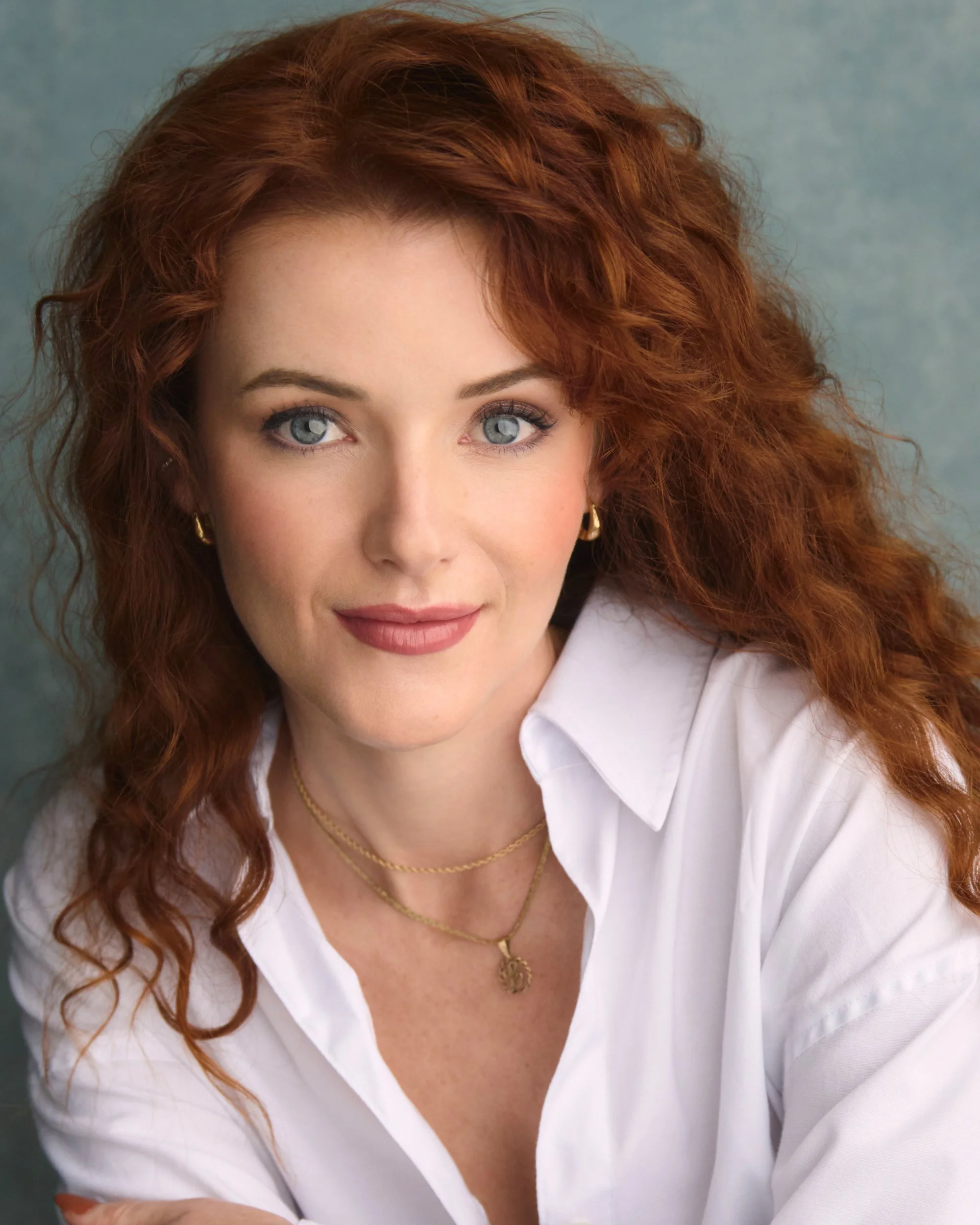 Close-up of a woman with curly red hair and blue eyes, wearing a white blouse, gold jewelry, and earrings, against a plain background.