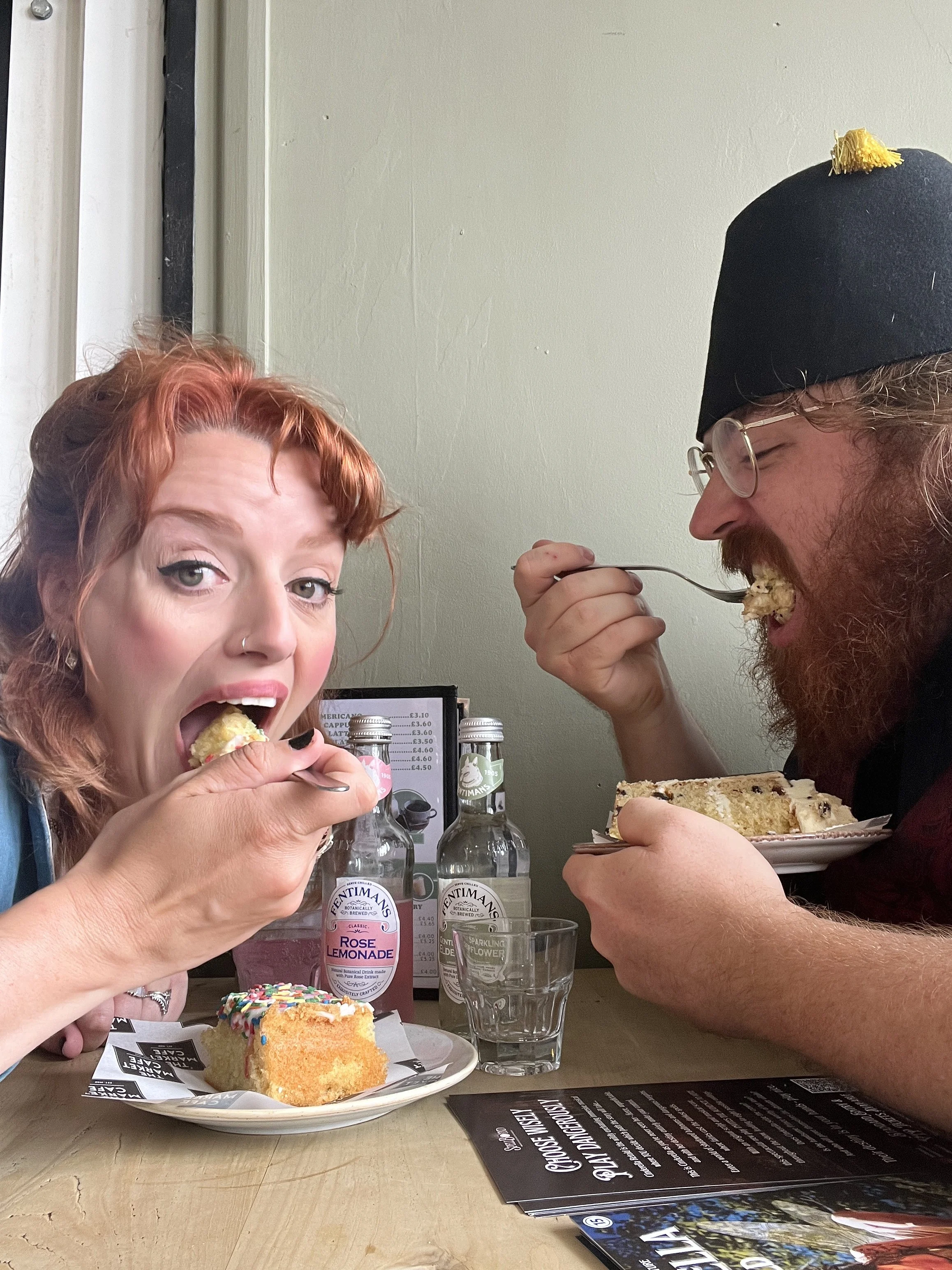 A woman with red hair and a man with glasses and a black hat are eating cake in a cafe. The woman is holding a fork and a piece of sponge cake with colorful sprinkles. The man is biting into a large slice of cake with frosting. There are bottles of d