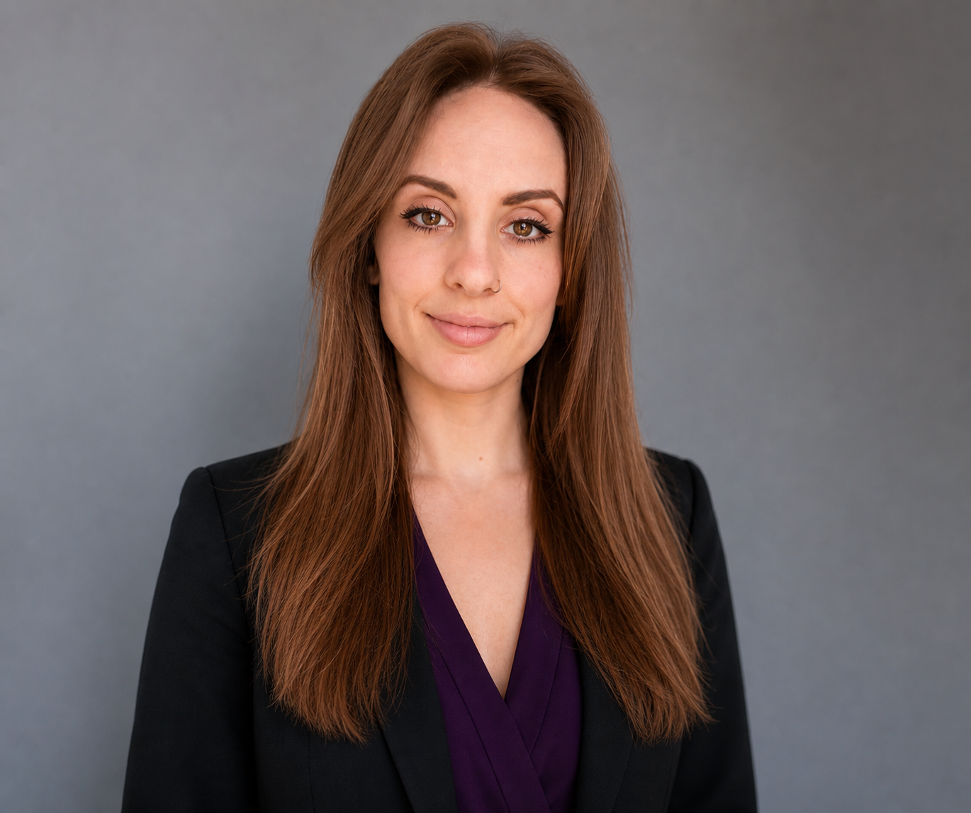 A portrait of a woman with long straight brown hair, light skin, wearing a black blazer and a purple top, standing against a plain gray background.
