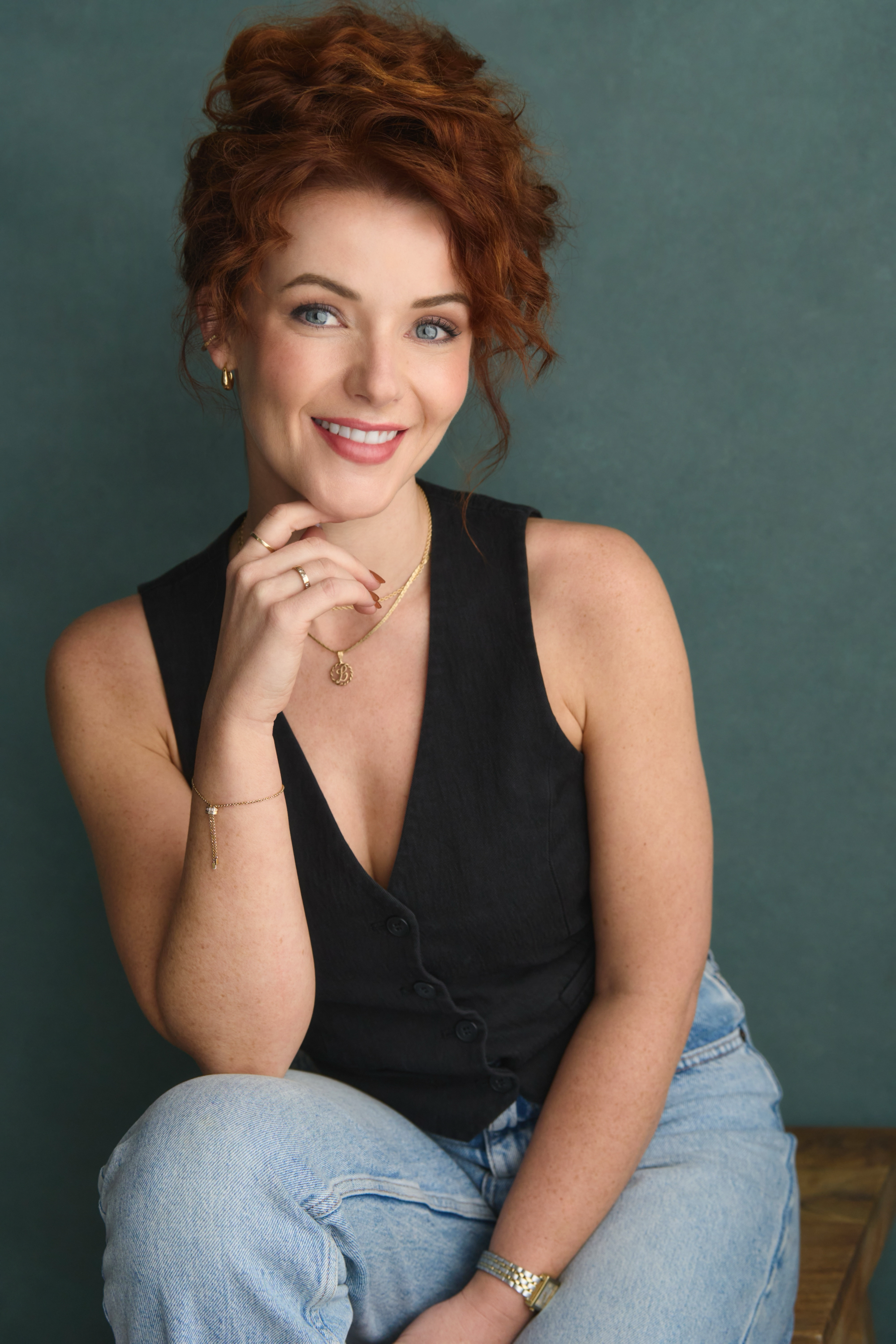 Young woman with curly red hair, wearing a black sleeveless top, sitting in front of a teal background, smiling and looking at the camera.