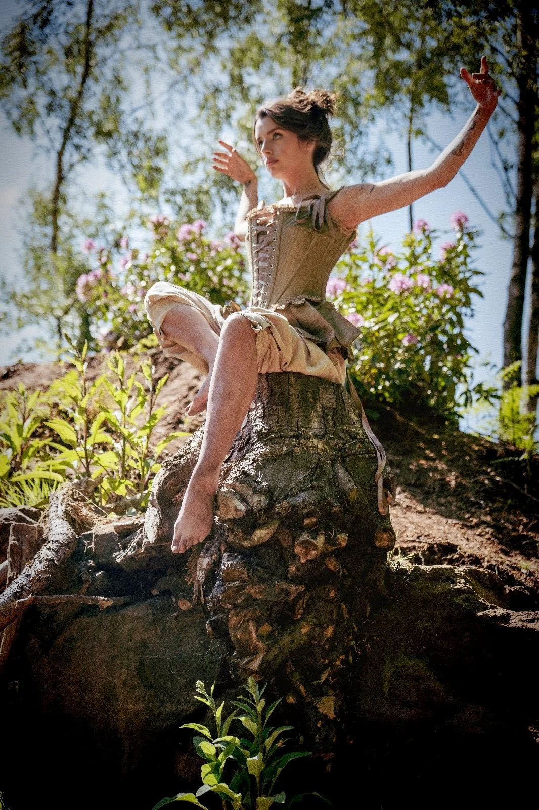 A woman in a beige, Victorian-style dress sits cross-legged on a large, fallen tree trunk in a forest, with her arms raised and surrounded by trees and pink flowers.