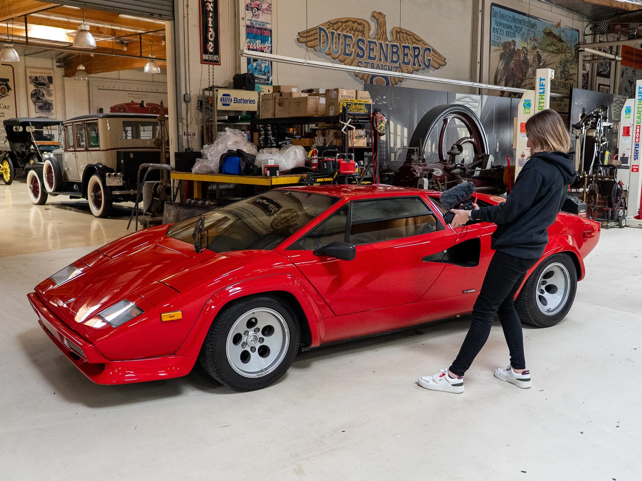A woman is filming a red sports car with a handheld camera in a garage filled with vintage cars and automotive memorabilia.