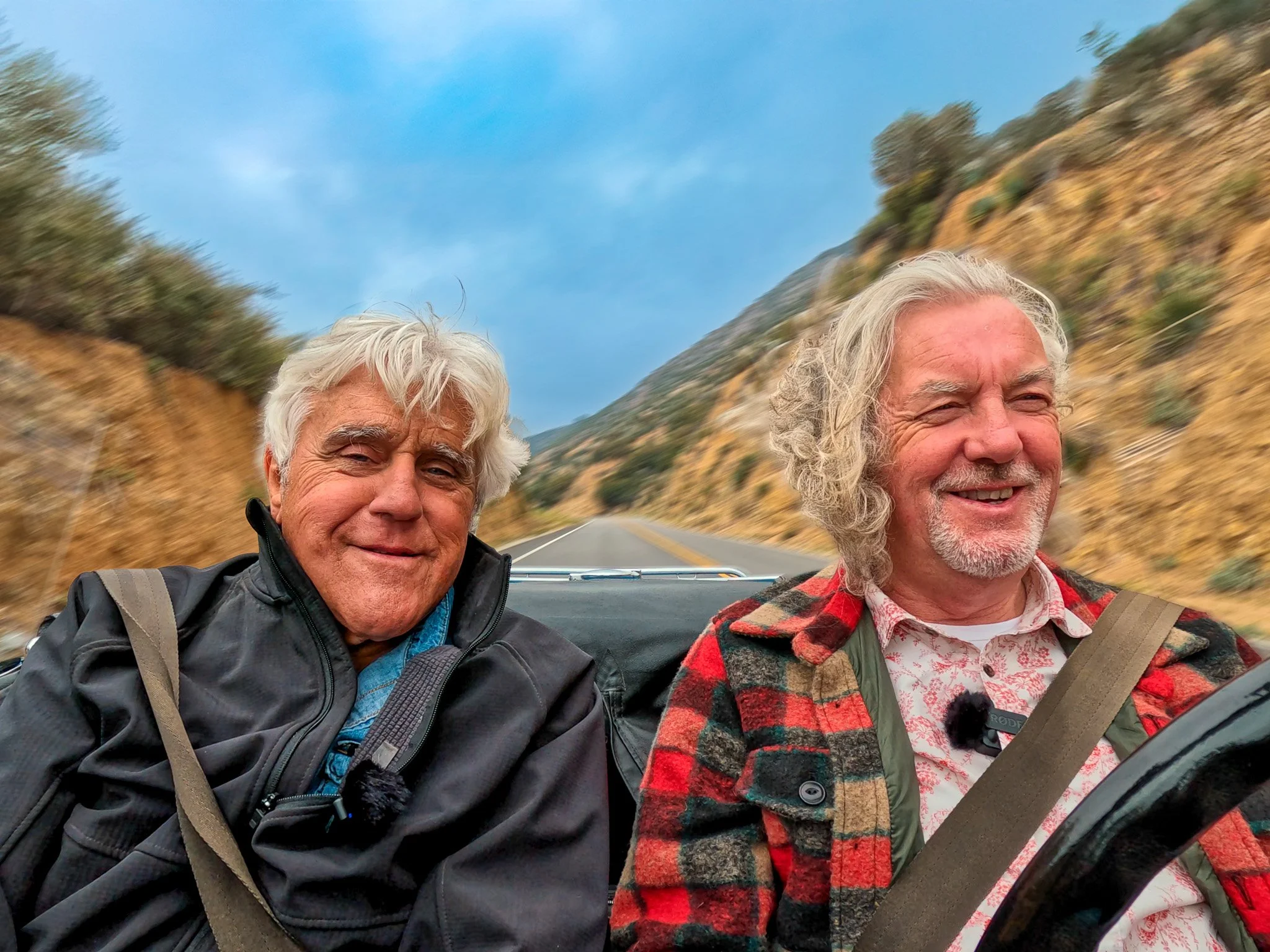 Two smiling older men riding in a convertible car on a mountain road with rocky hills and sparse vegetation, one wearing a black jacket and the other wearing a plaid jacket and patterned shirt.