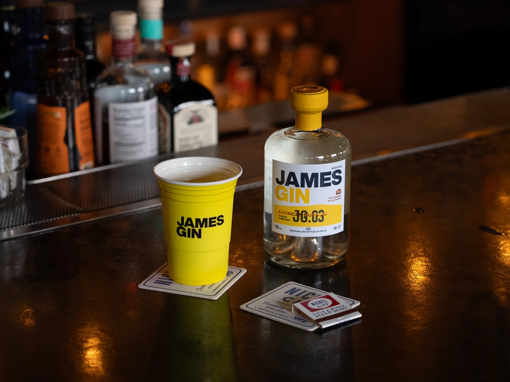 A bottle of James Gin and a yellow cup labeled James Gin on a bar counter, with a coaster underneath the cup and playing cards beside them.