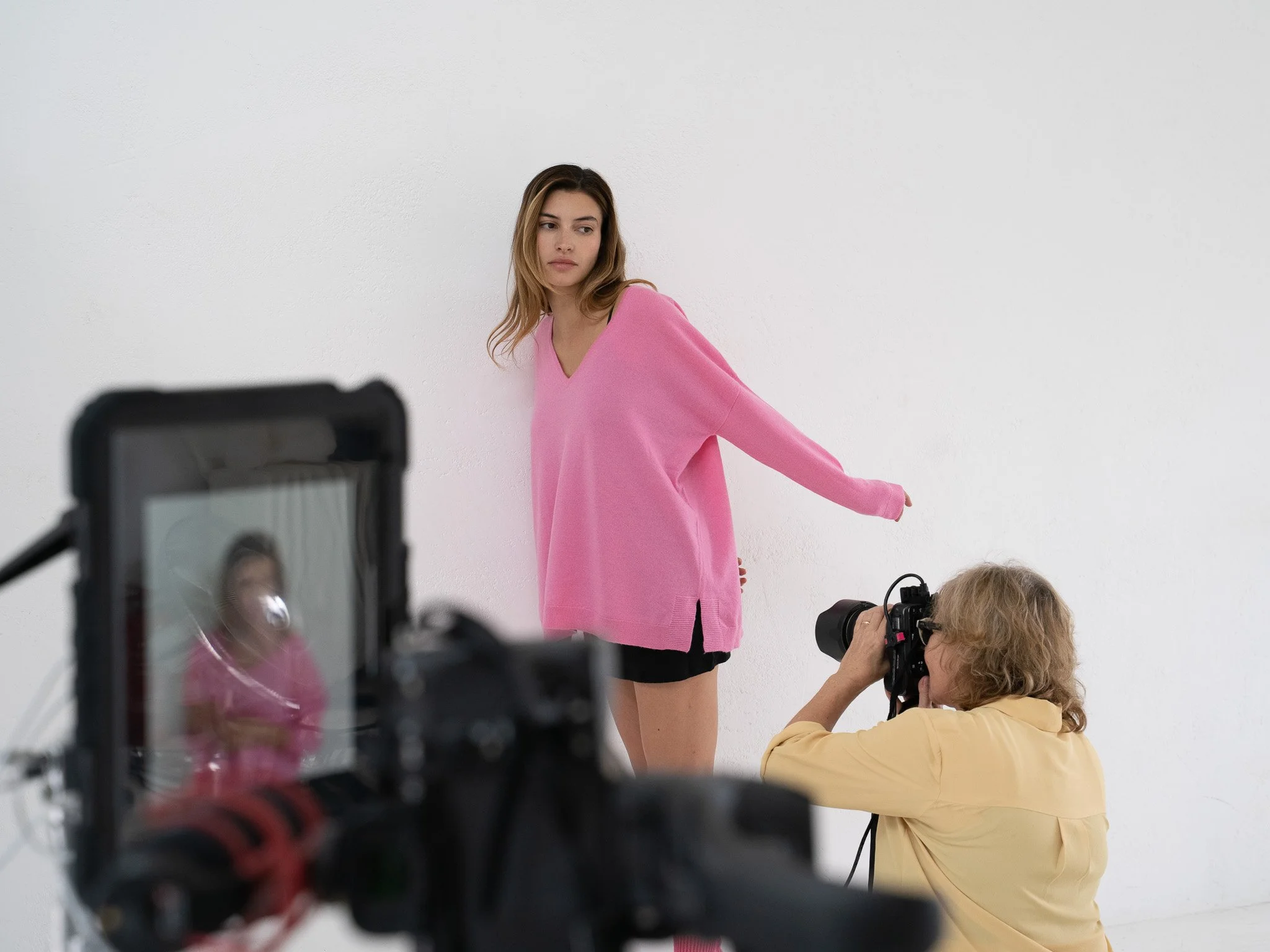 A woman in a baker miller pink sweater poses for a photoshoot against a white wall while a photographer takes her picture, and a third person captures the scene with a camera.