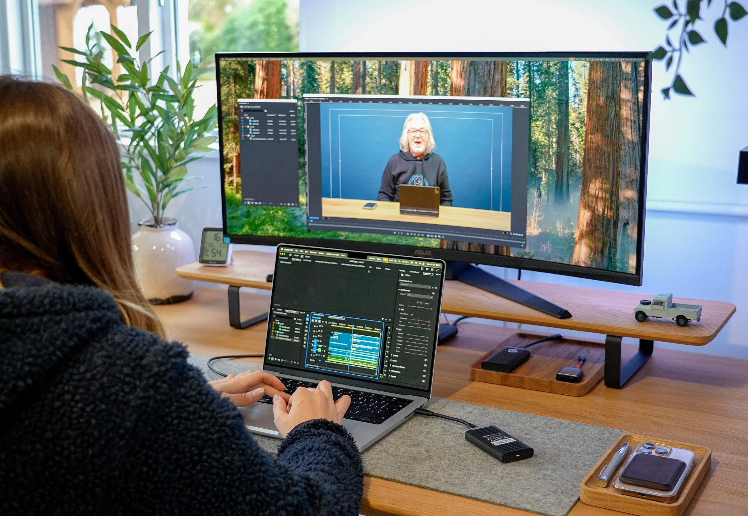 Person working on a laptop at a desk with a large monitor displaying a video conference, a small plant, a digital clock, and other office items.
