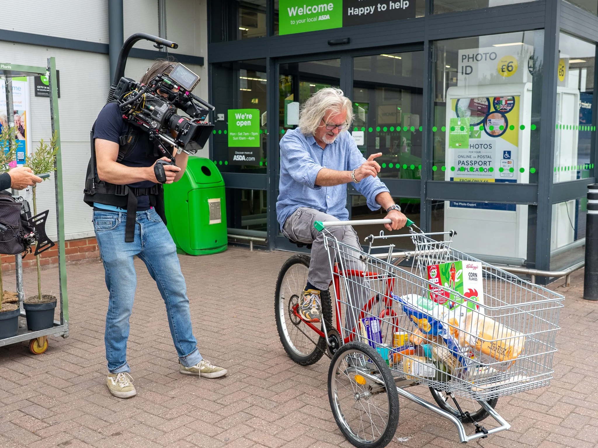 James May riding a bicycle with a shopping cart filled with grocery bags outside a grocery store. A cameraman is filming him, and another person is partially visible. Filming taking place for James May's Shedload of Ideas TV show for Quest.