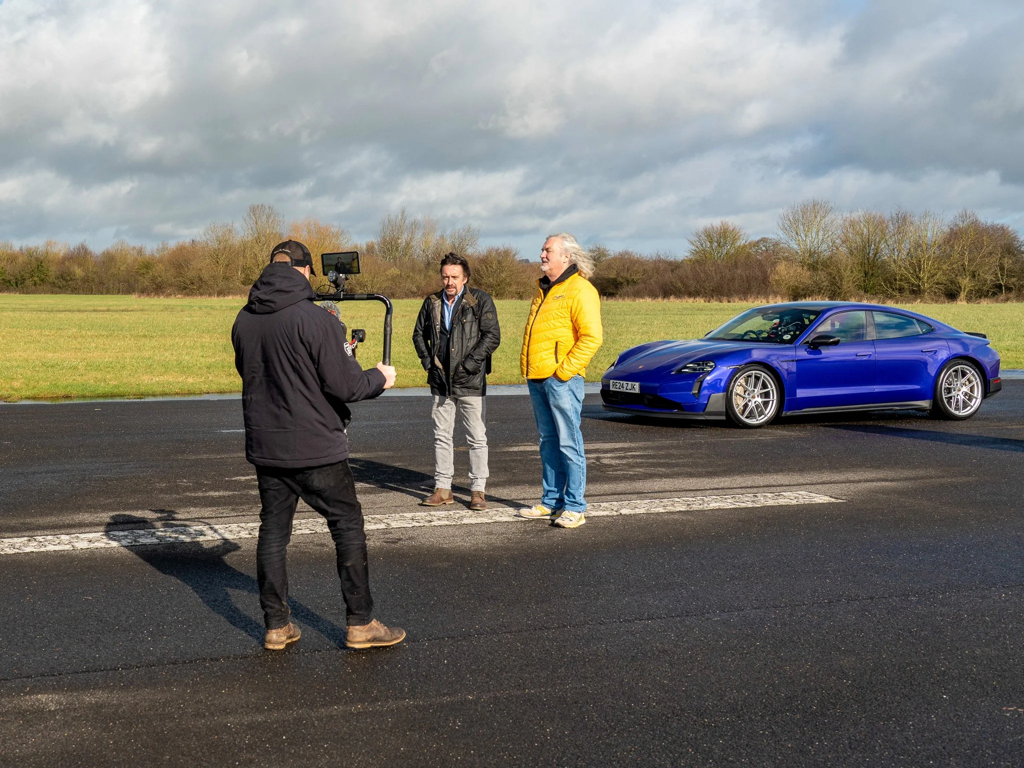 Three men being filmed next to a blue sports car on an open road with a grassy field and trees in the background under cloudy skies.