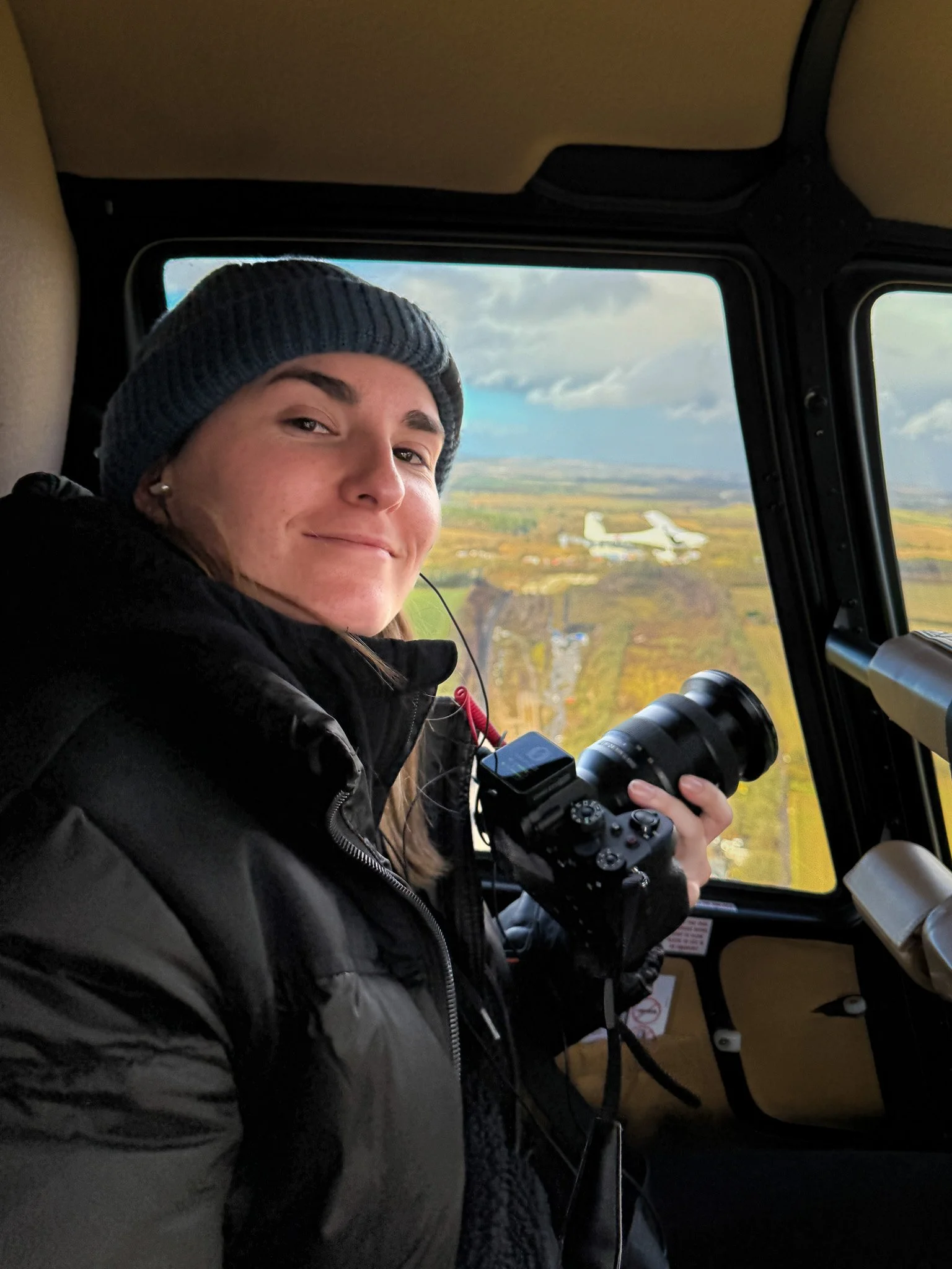 Woman in a beanie and black jacket inside a helicopter, holding a camera, with landscape and airplane visible through the window.