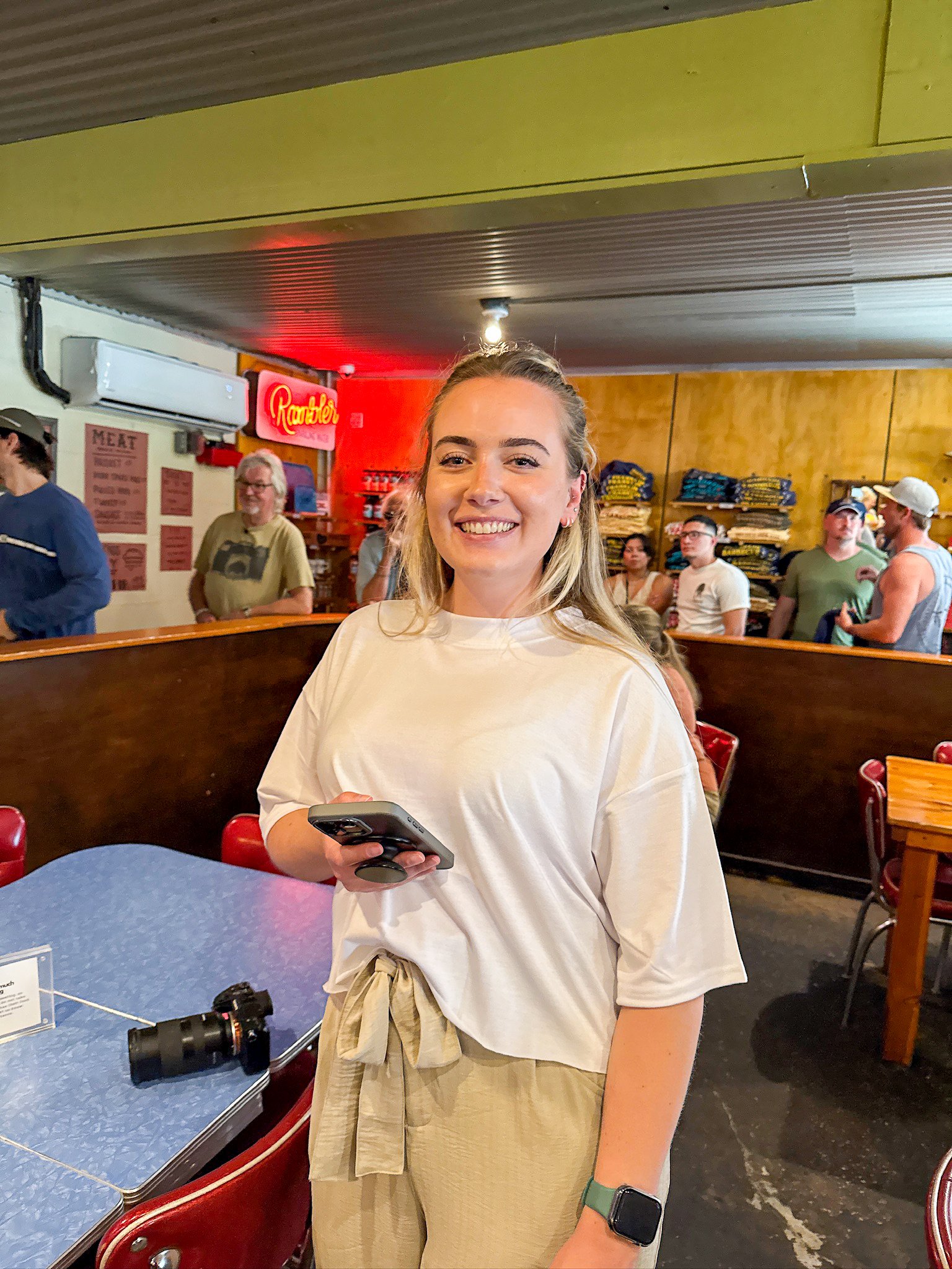A young woman smiling while holding a phone in a casual restaurant or diner setting. She is wearing a white t-shirt, beige pants, a watch, and has blonde hair. There are other people in the background and a camera on the table.