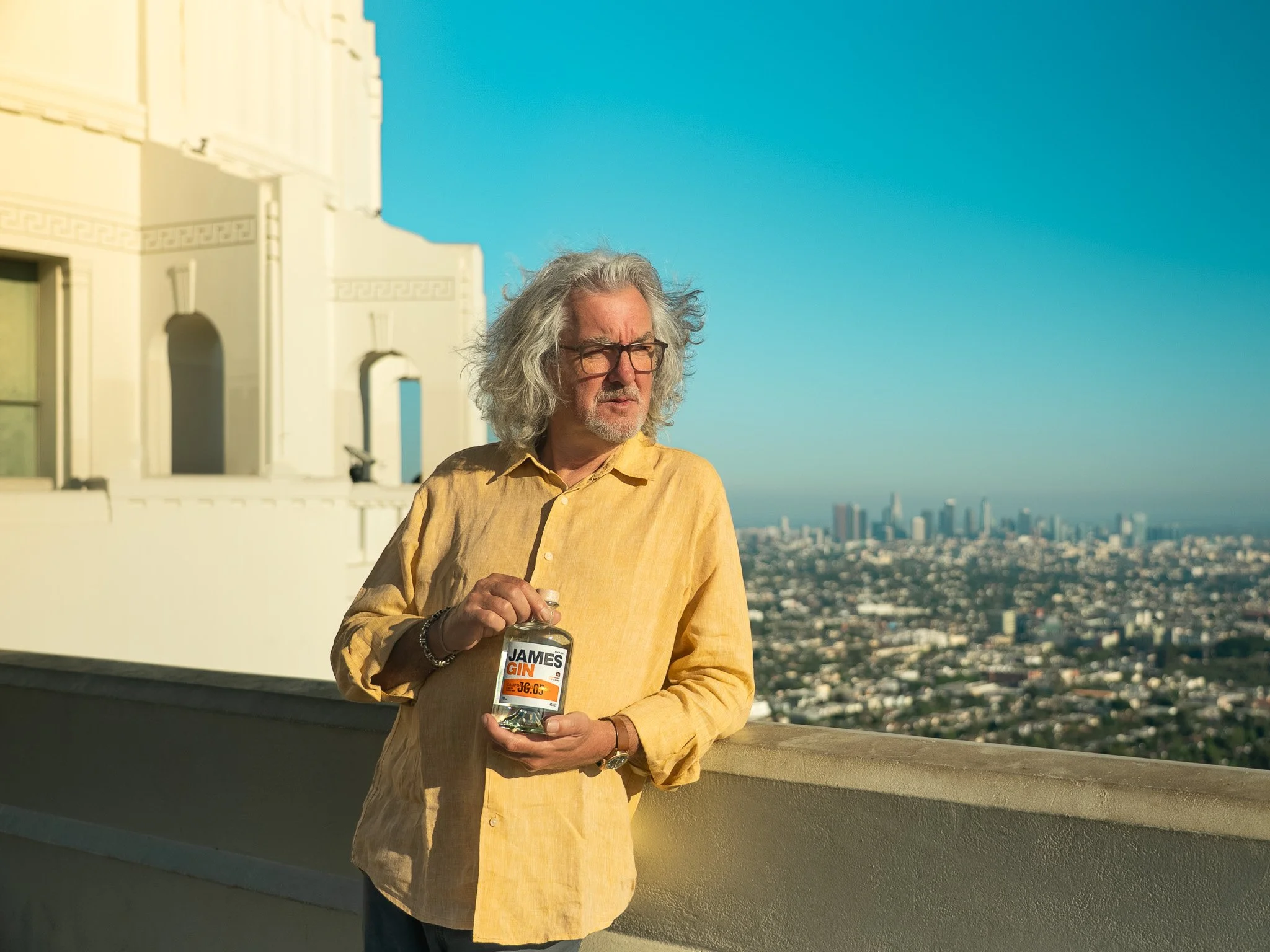 James May stands on a rooftop balcony overlooking Los Angeles with tall buildings and a clear blue sky. He holds a bottle labeled 'James GIN' in his hands.