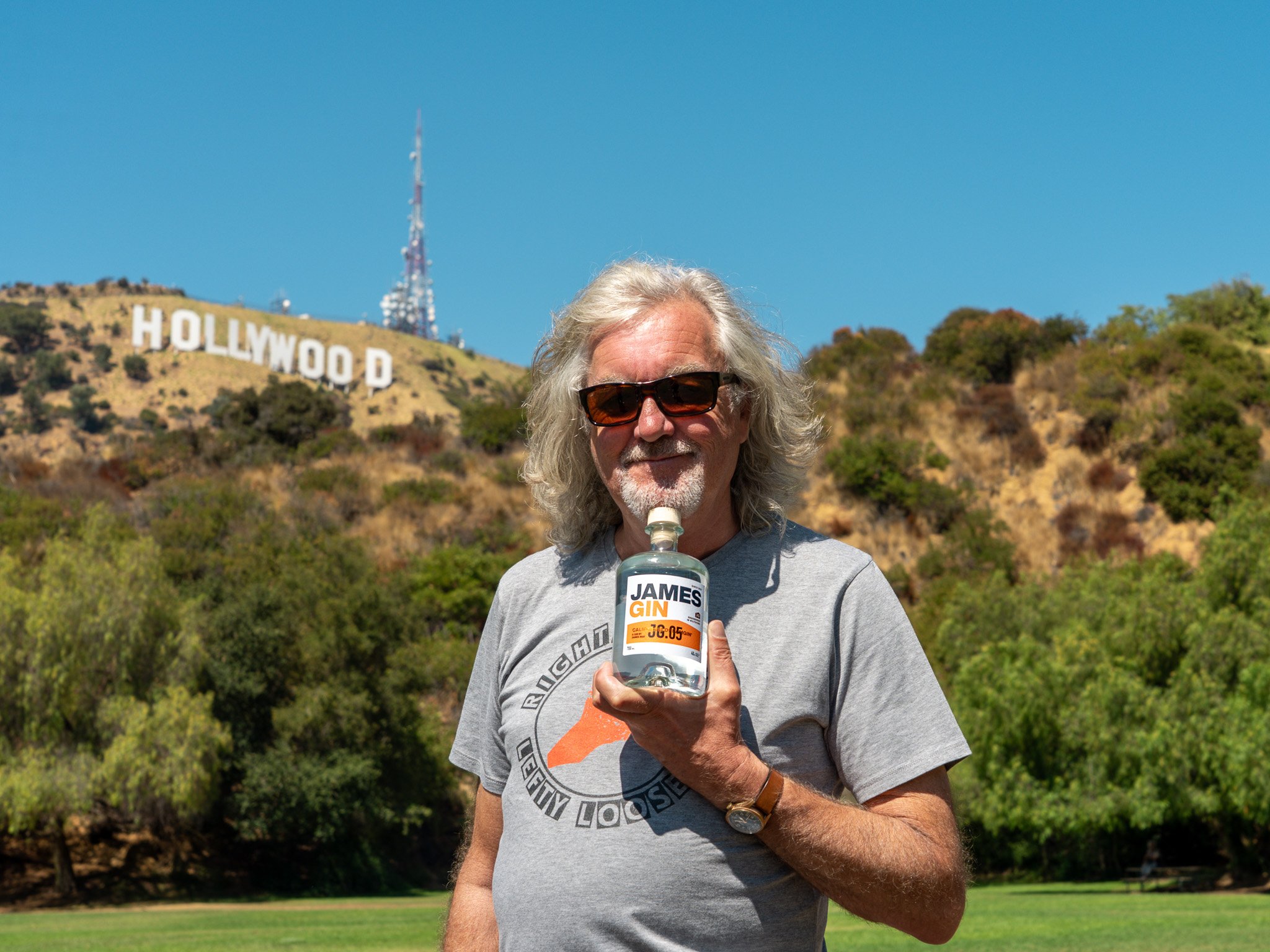 An elderly man with gray hair and beard, wearing sunglasses and a gray t-shirt, holding a bottle of James Gin, standing outdoors with the Hollywood sign on the hillside behind him and a clear blue sky.
