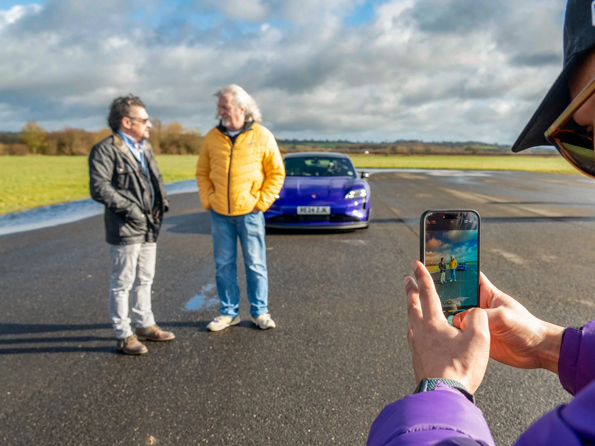 James May and Richard Hammond standing and talking in the middle of an open road, with a blue sports car behind them. Another person in a purple jacket is taking a photo of the men with a smartphone, partially visible on the right side of the image.