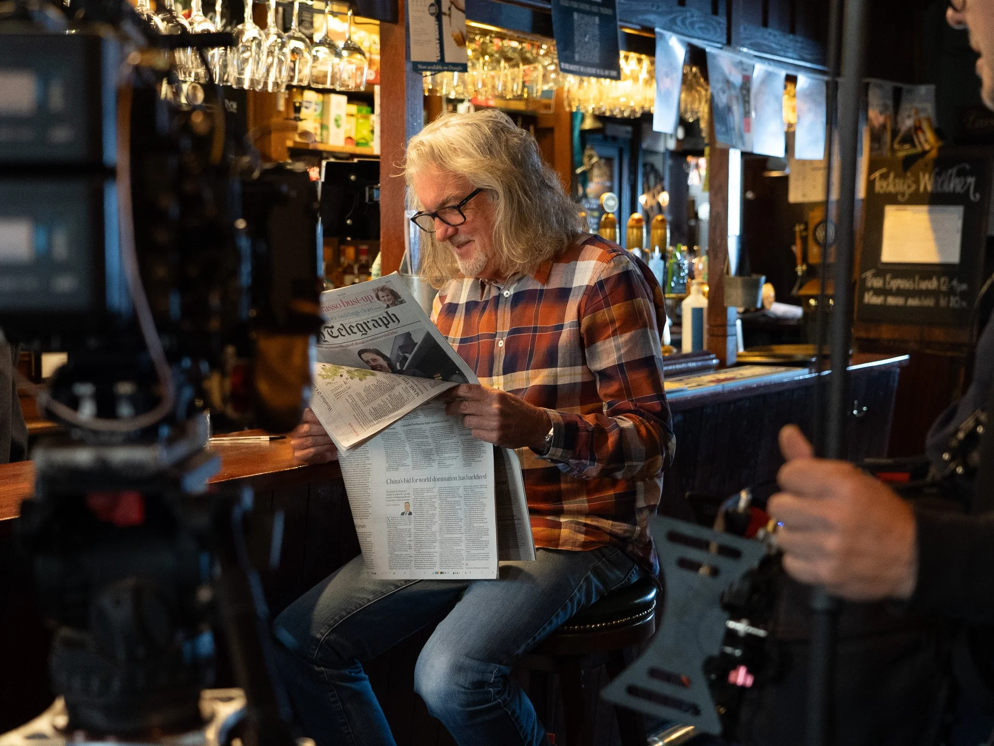A man with long gray hair and glasses reading a newspaper at a bar, surrounded by filming equipment.