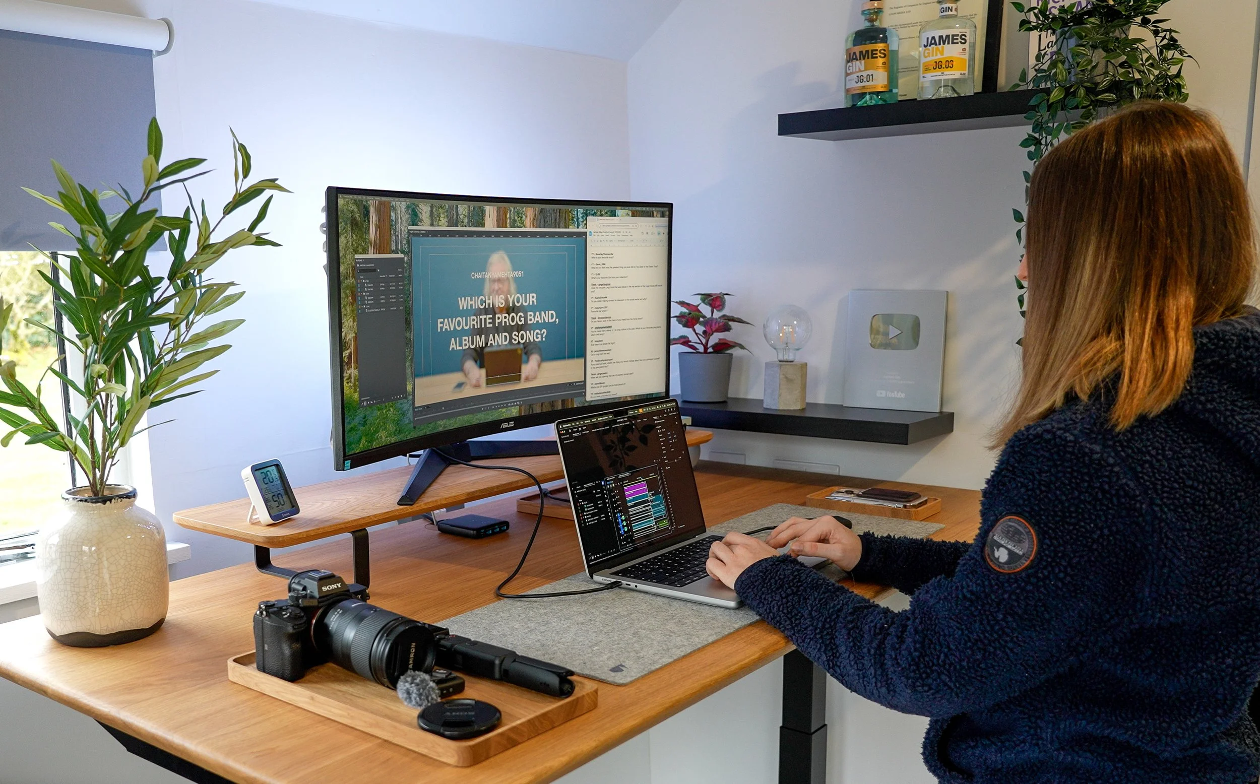 A woman with brown hair working at a wooden desk with a laptop and a large monitor. The monitor displays a presentation slide asking about favorite music, album, and song. The desk has a potted plant, a digital clock, a camera, and various accessories. On the wall, there are shelves with bottles of gin, a YouTube play button award, and a small plant. A window provides natural light.