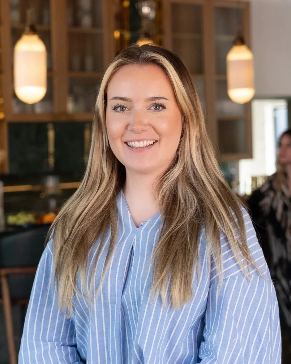 Eve Rawlings, a smiling woman with long blonde hair and blue eyes, wearing a blue and white striped shirt, standing in a modern restaurant or café with warm lighting and wooden decor.