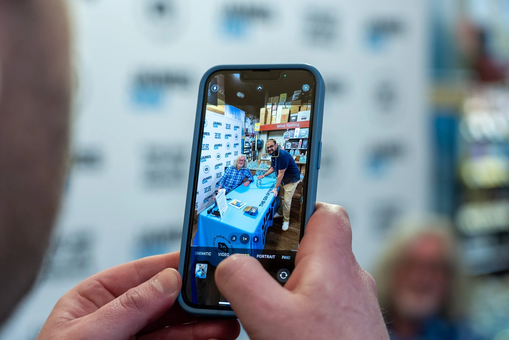 Person taking a photo with a smartphone of two men at a table during a book signing event at a bookstore, with shelves of books and a sign that says "Wine Tasting" in the background.