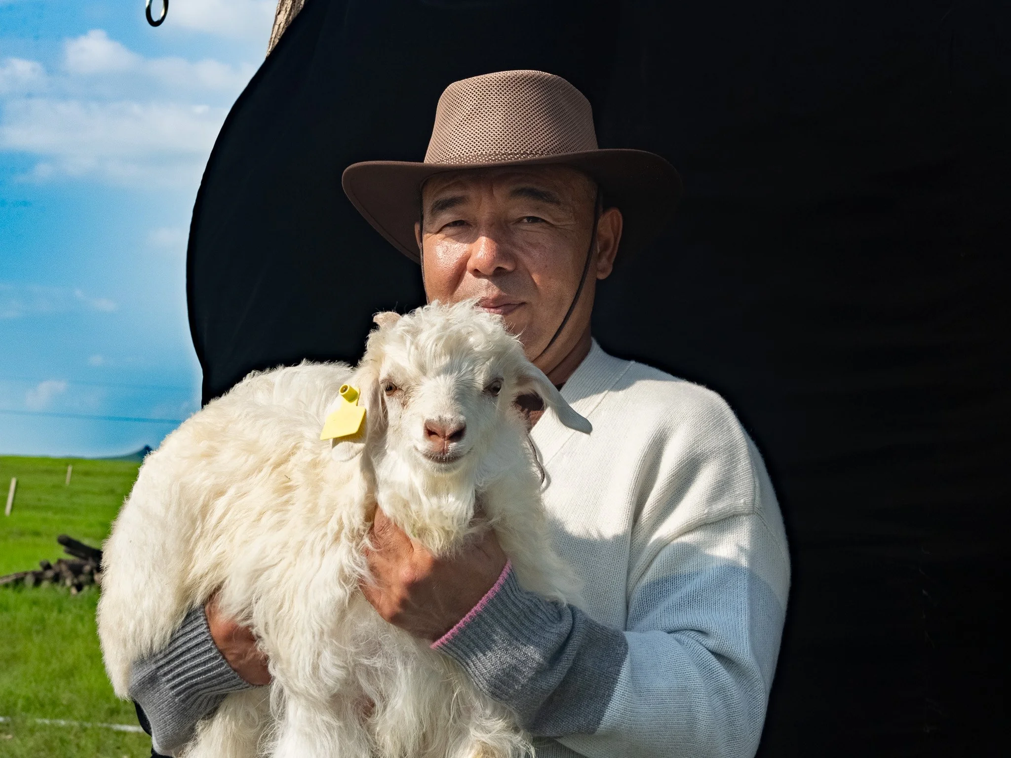 A man wearing a brown hat and a light-colored sweater holding a fluffy white baby goat in front of a black background, outdoors with a grassy field and blue sky in the distance.