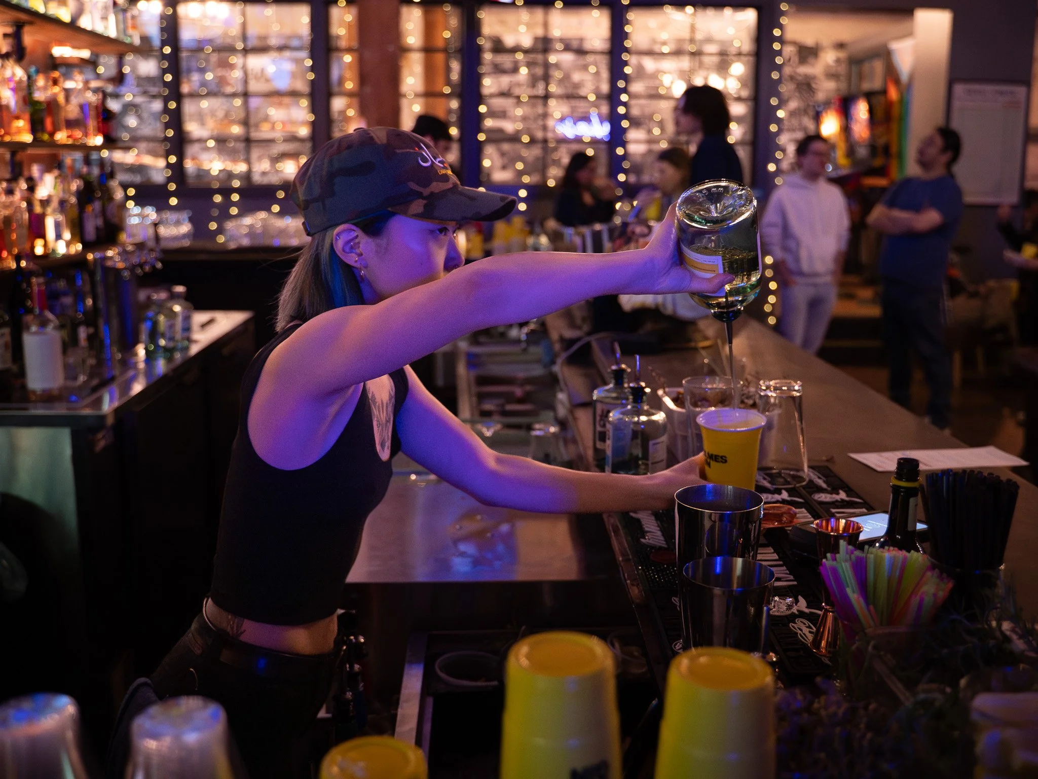 A female bartender pouring a drink from a glass bottle into a glass at a bar with dim lighting and string lights in the background.