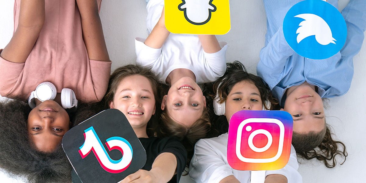 Five children lying on the floor looking up, holding signs with social media logos TikTok, Instagram, Twitter, and Snapchat.