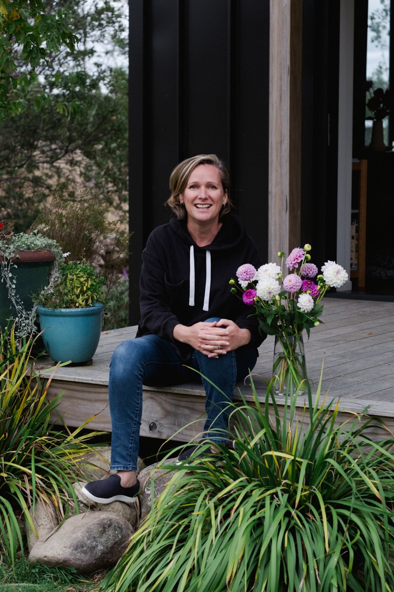 Woman sitting on the edge of a wooden deck surrounded by potted plants and a bouquet of pink and white flowers in a glass vase, with greenery and part of a modern black house in the background.