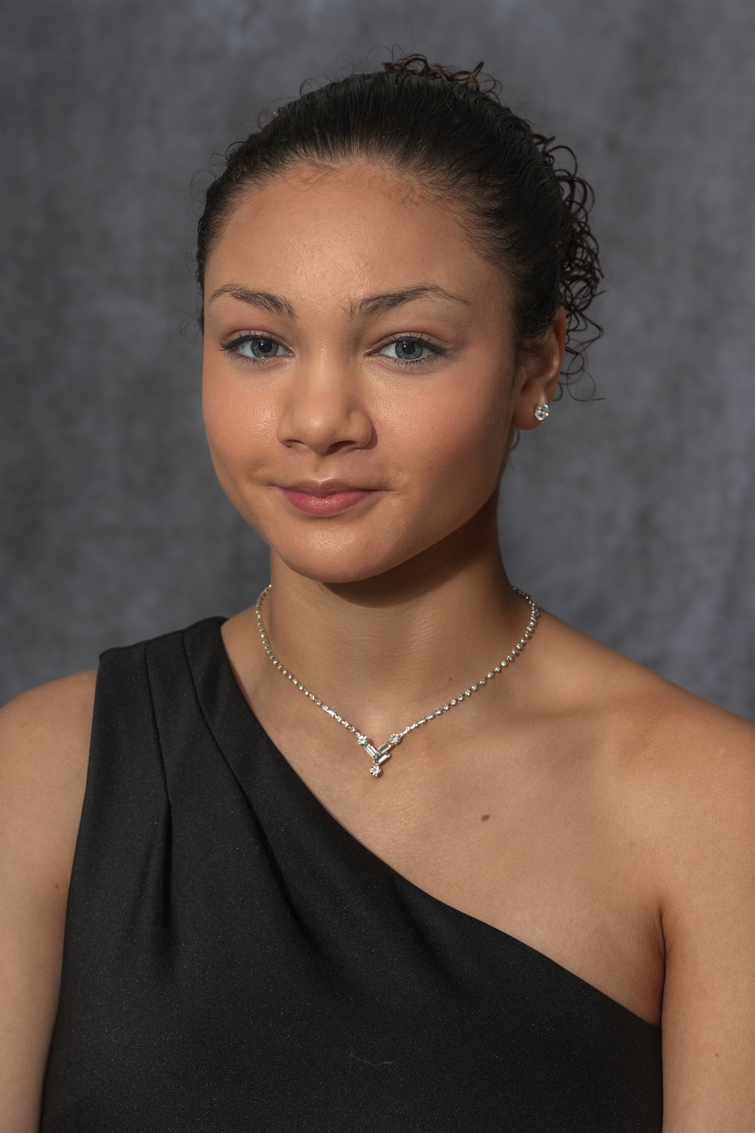 A young woman with curly hair tied back, wearing a black sleeveless dress, a necklace, and earrings, standing against a gray background.