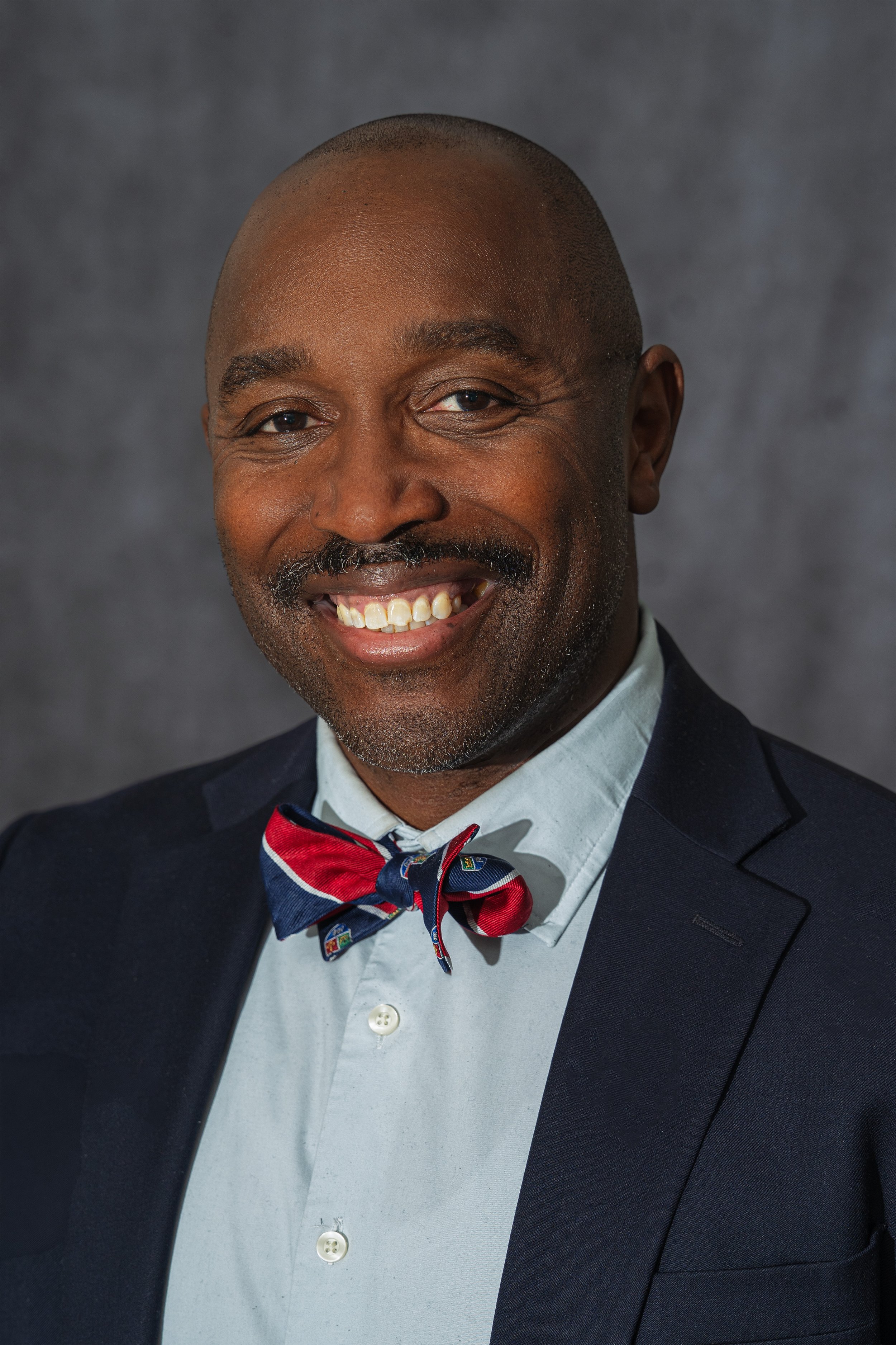 Portrait of a smiling man with a mustache and goatee, wearing a navy suit, light blue shirt, and a red, blue, and white bow tie.
