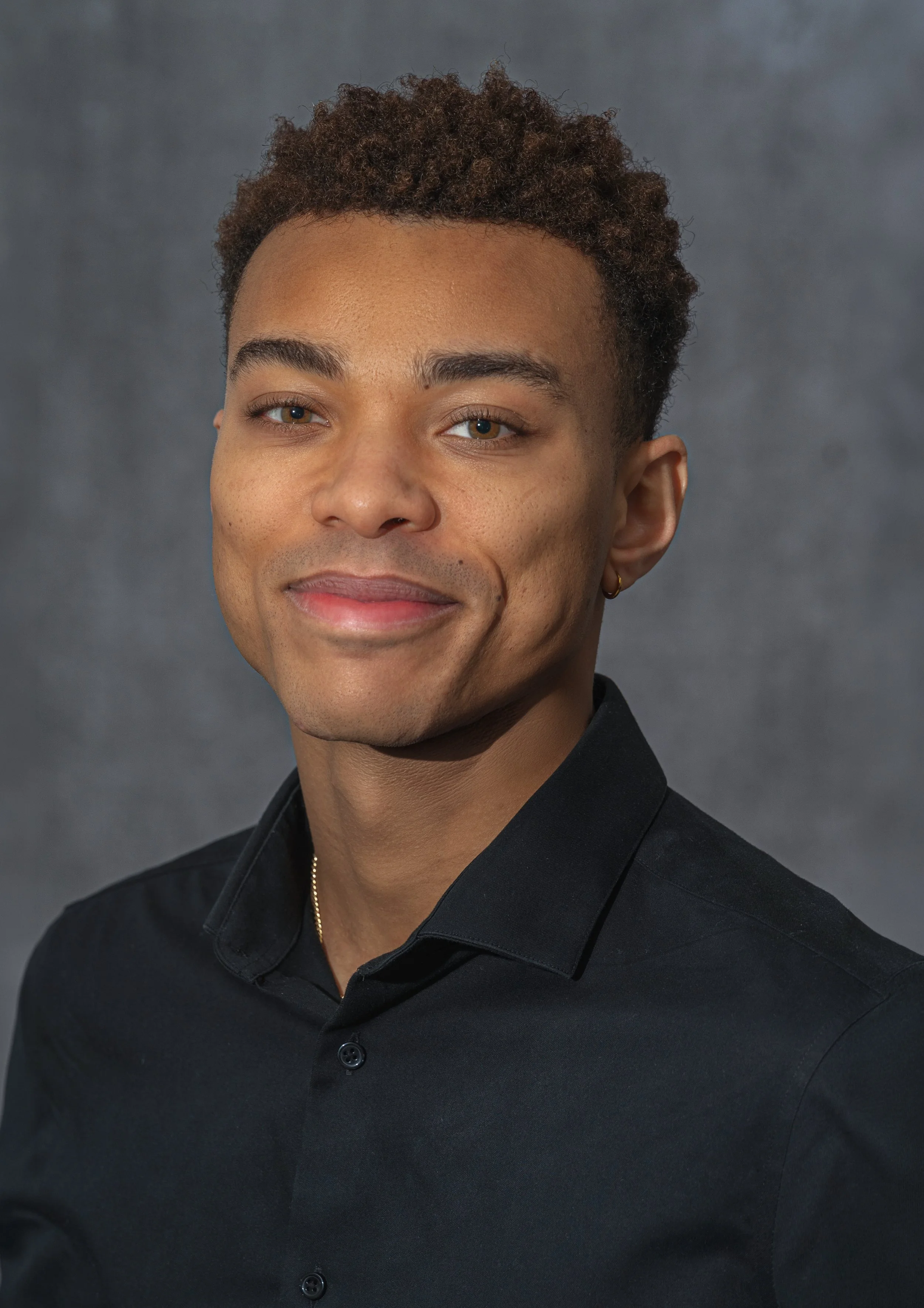 A young man with short curly hair, wearing a black shirt, smiling, against a gray background.