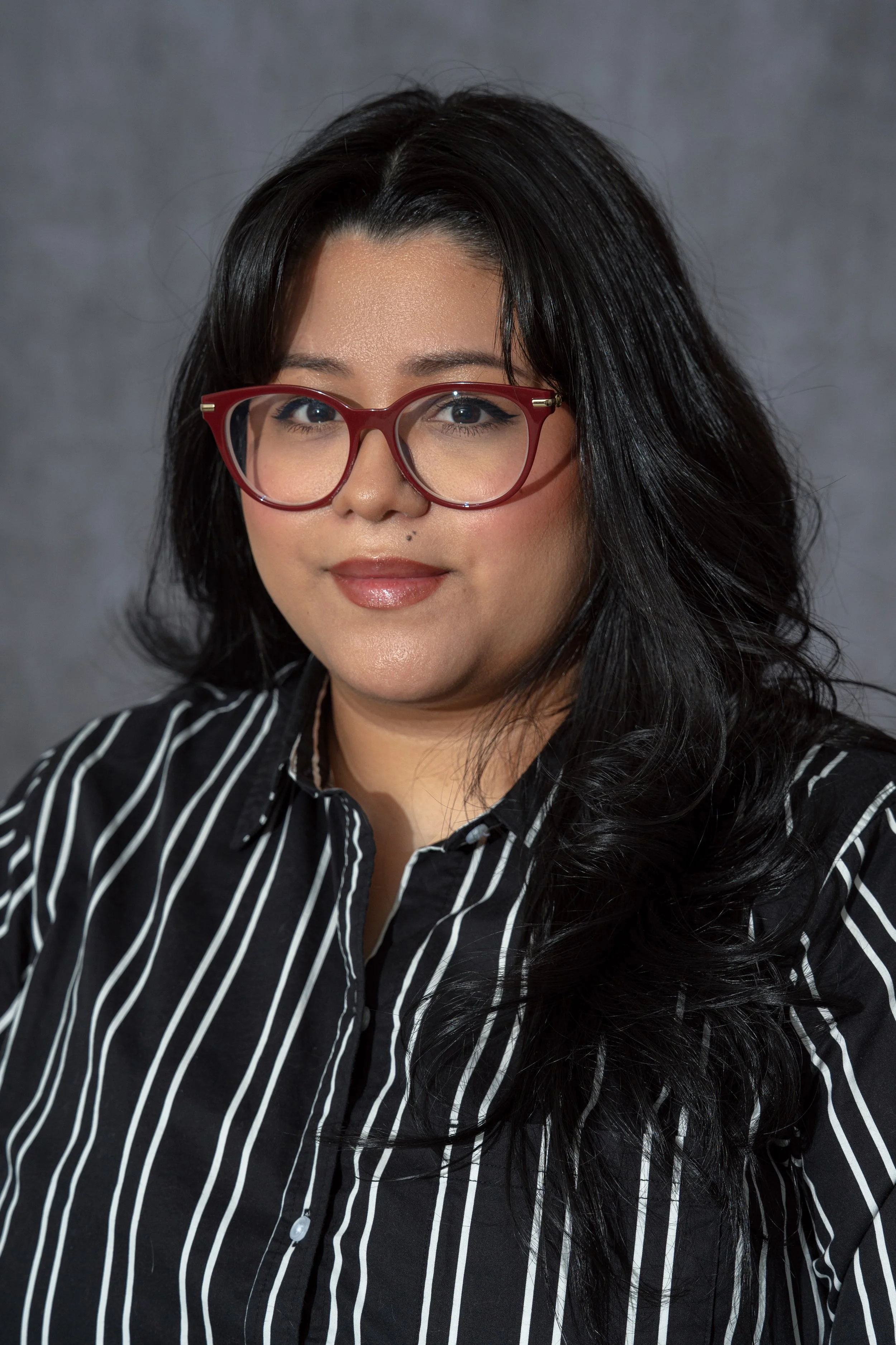 A woman with long black hair wearing red glasses and a black and white striped shirt, posed against a gray background.
