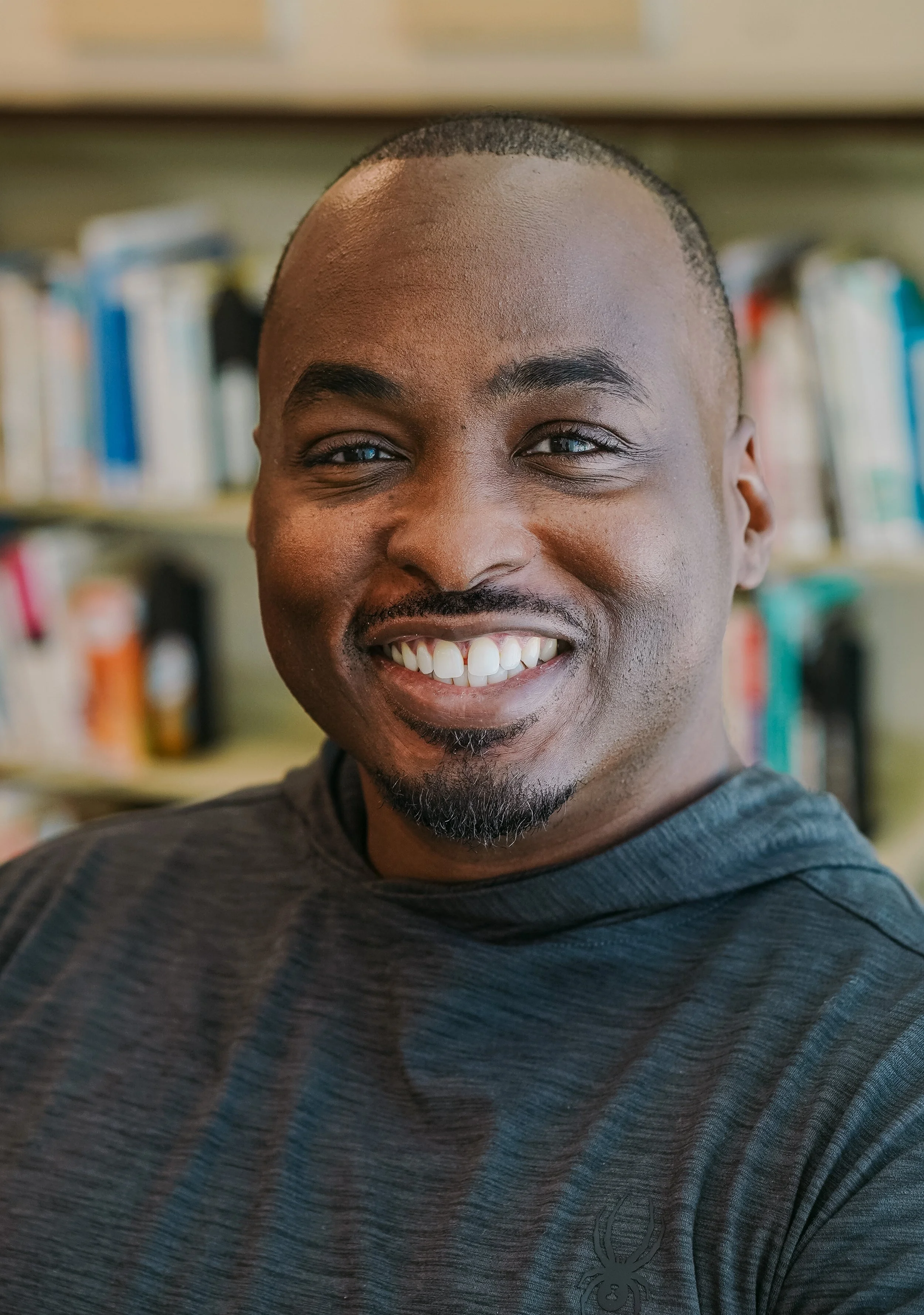 A smiling man with a goatee and short hair in a library, wearing a dark gray athletic shirt.