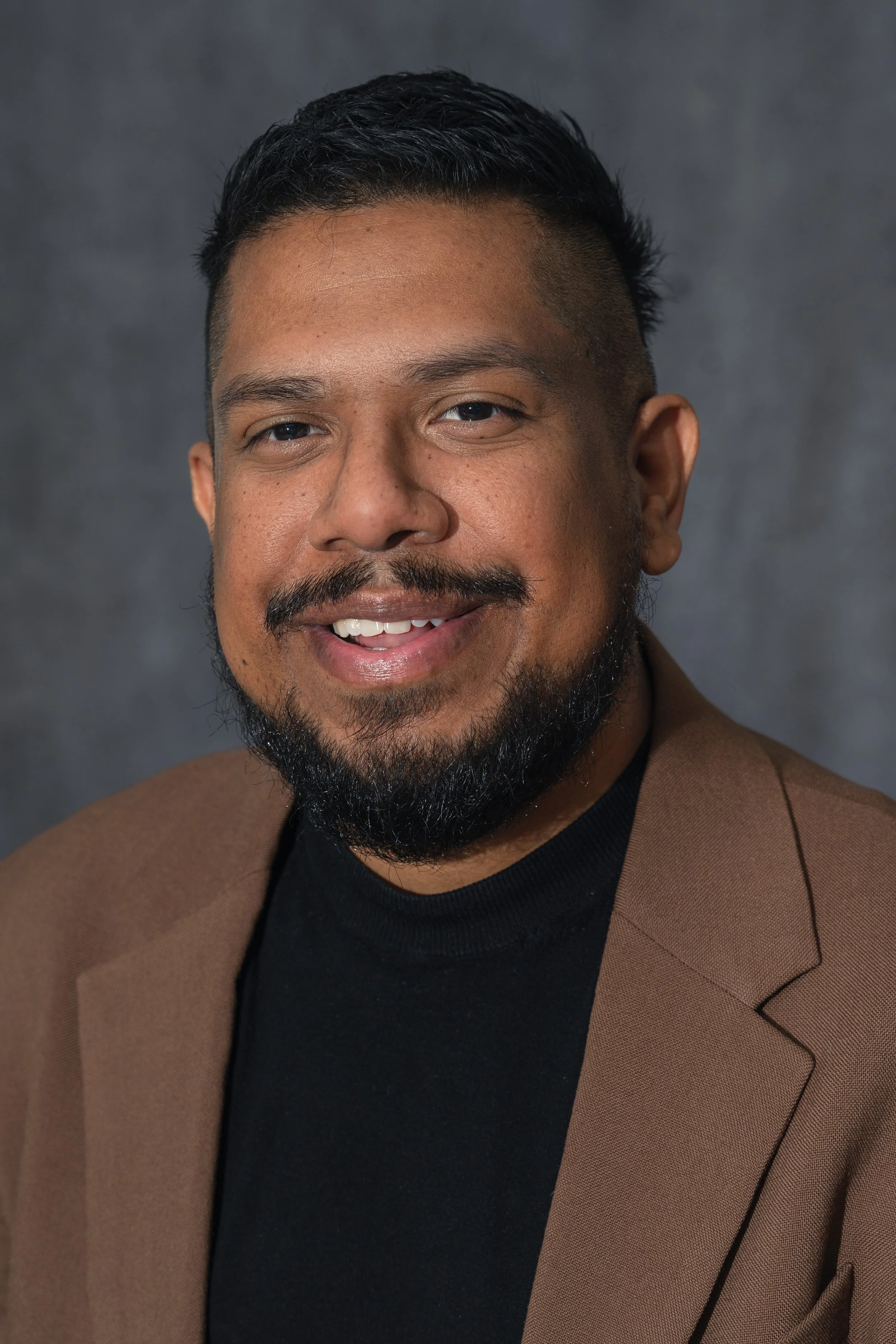 Portrait of a smiling man with a beard and mustache, wearing a black shirt and a brown blazer against a gray background.