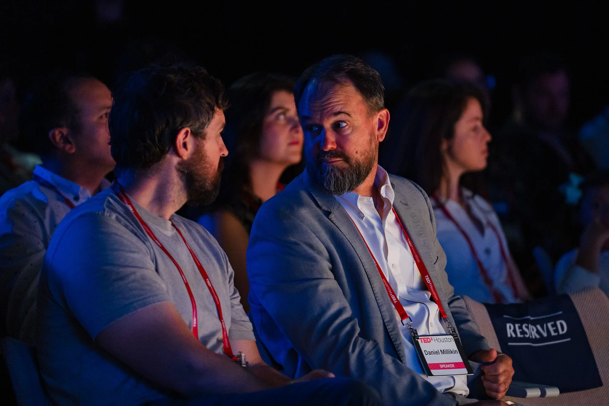 Two TEDxHouston attendees sit in the audience and share a look of surprise and intrigue during a speaker presentation.