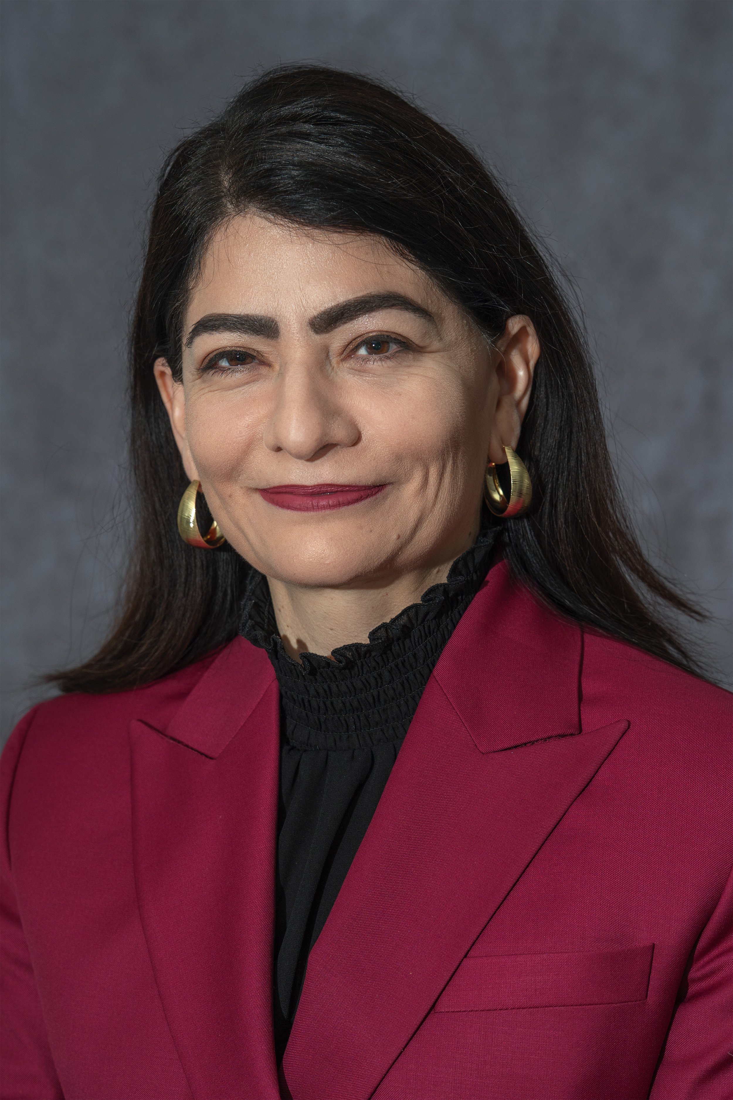 A woman with dark hair, wearing gold hoop earrings, a black ruffled blouse, and a burgundy blazer, smiling slightly against a gray background.