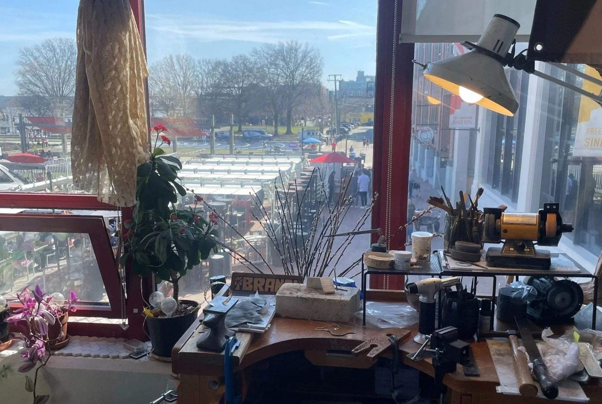 View from inside a workshop overlooking a street with outdoor market stalls, trees, and buildings in the background. Sunlight streams through large window.